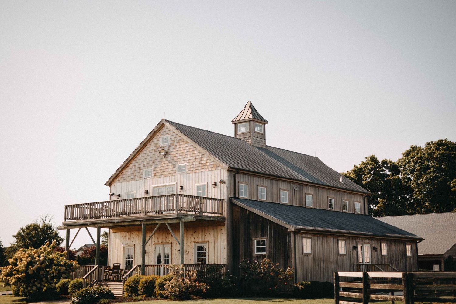 Rustic wooden barn wedding venue with cupola and wraparound balcony under clear sky.