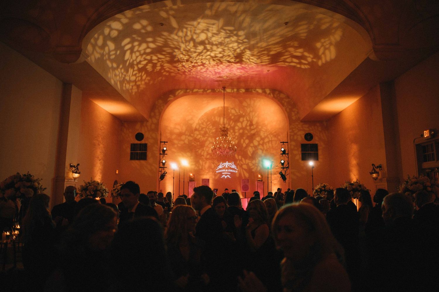 Guests dance at romantic wedding reception with warm amber lighting and floral ceiling projections.