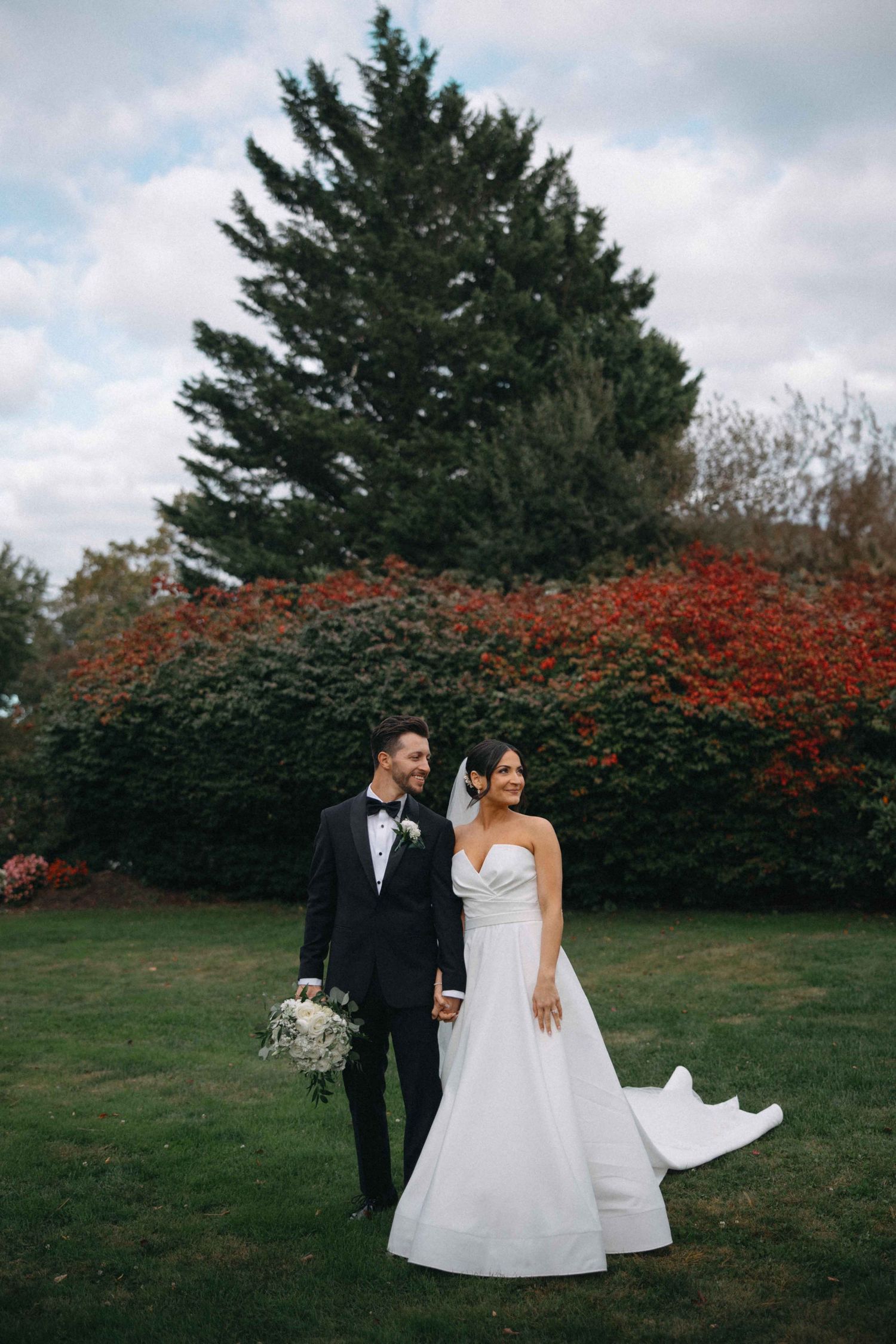 Bride and groom walking hand in hand in a garden with colorful autumn foliage behind them.