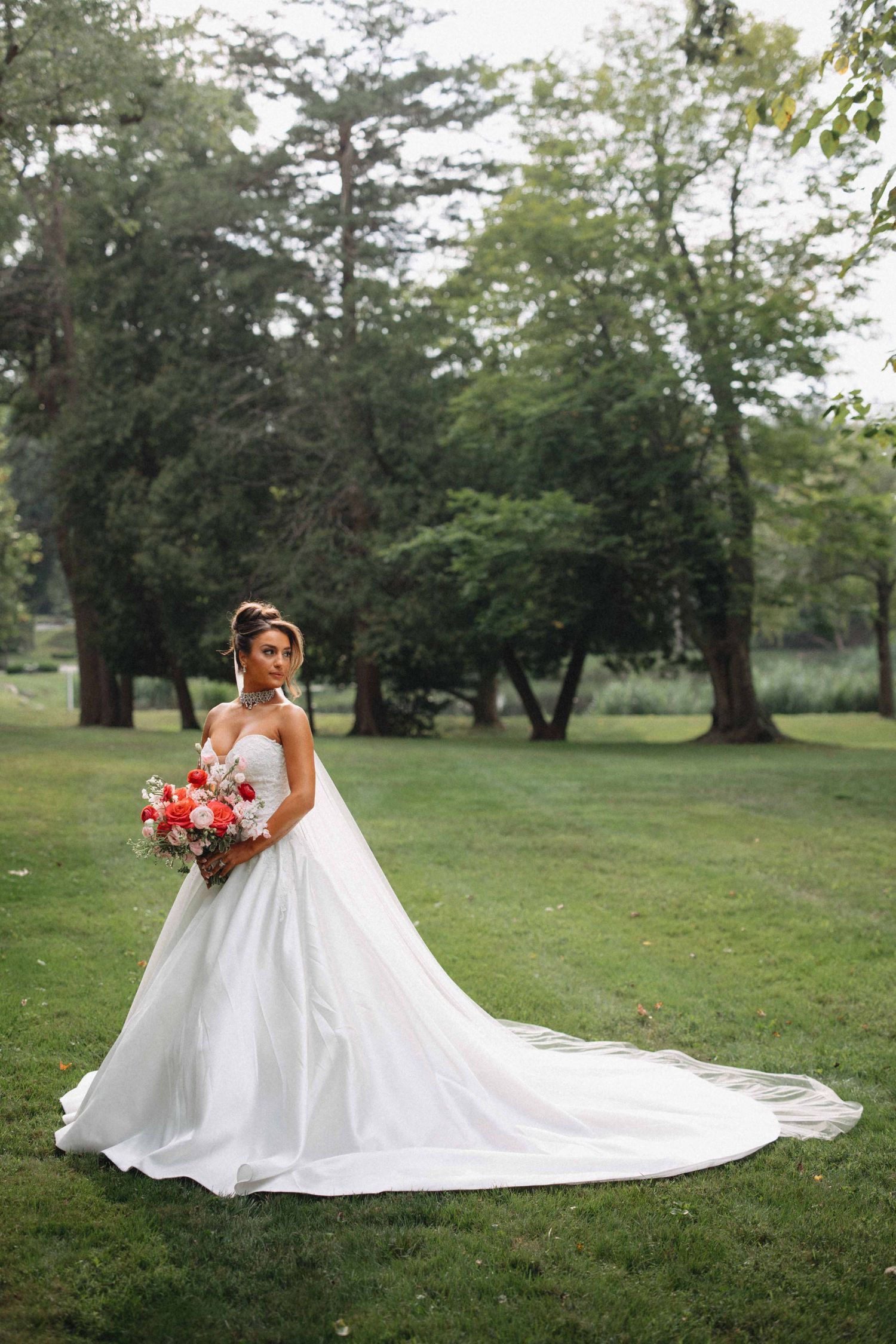 Elegant bride in a flowing white ball gown with long train holds a red bouquet in a lush green park.