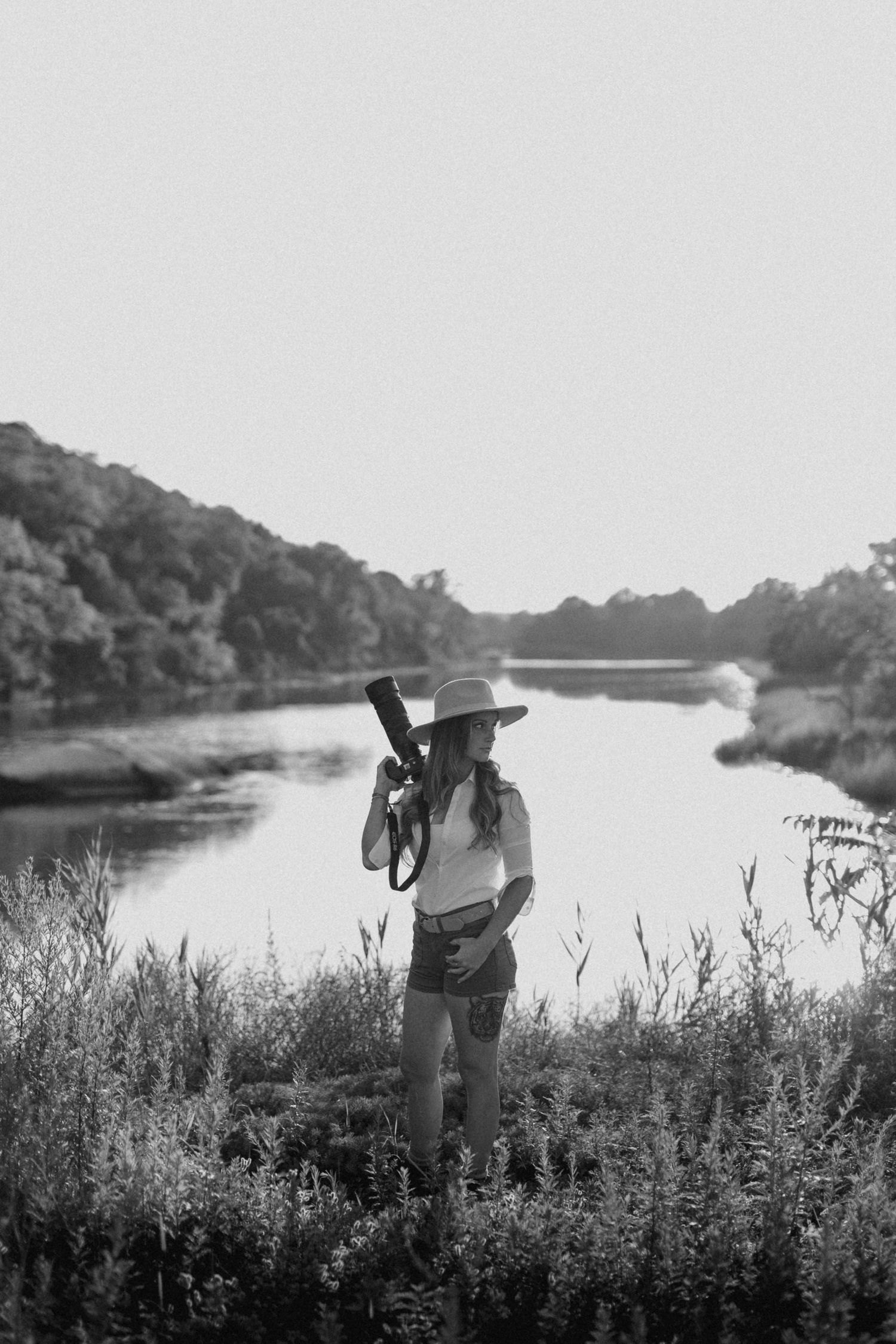 Black and white photo of a female photographer holding a camera by a serene river surrounded by lush greenery.