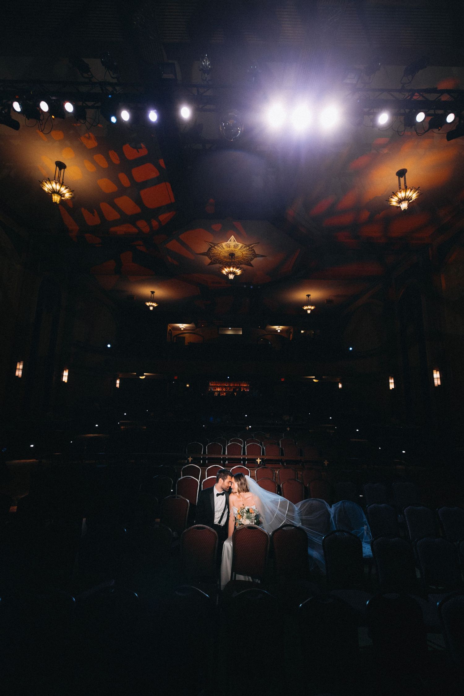 Bride and groom share a romantic moment in the center of a grand, dimly lit theater.