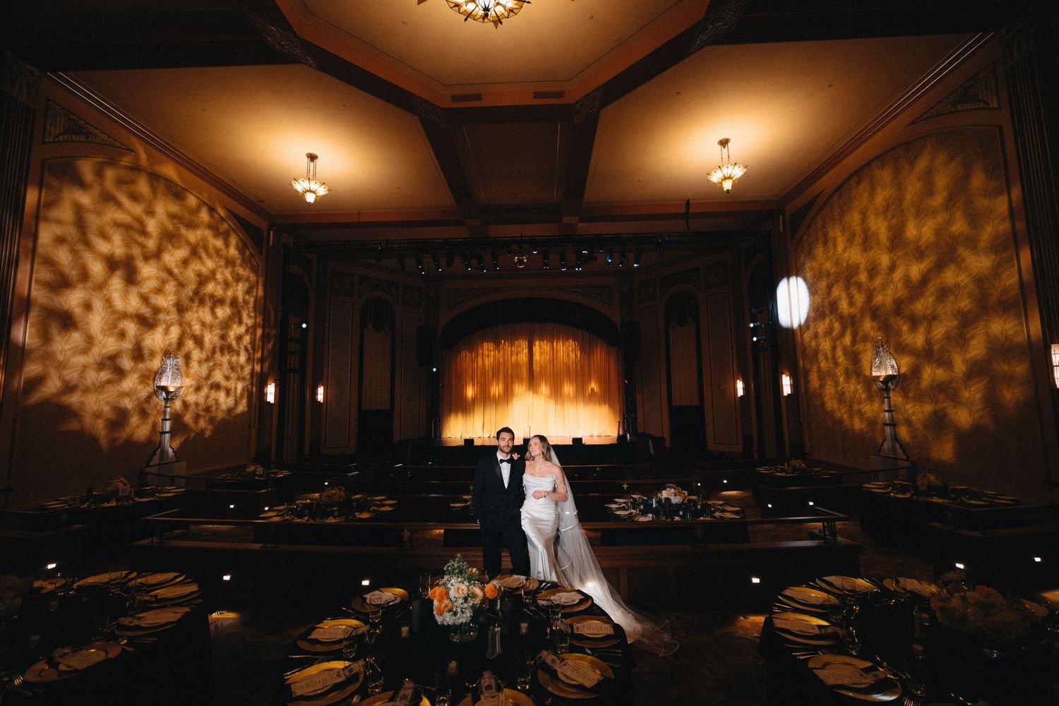 Bride and groom stand together in an elegantly lit wedding reception hall with golden drapery and chandelier lighting.