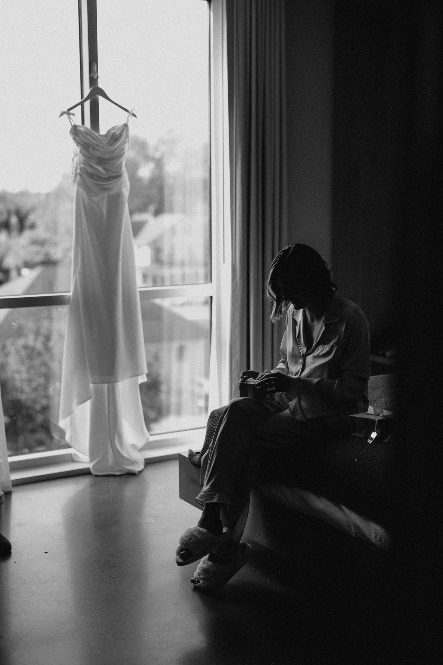 Bride sitting by window in robe reviewing photos beside hanging wedding dress in black and white.