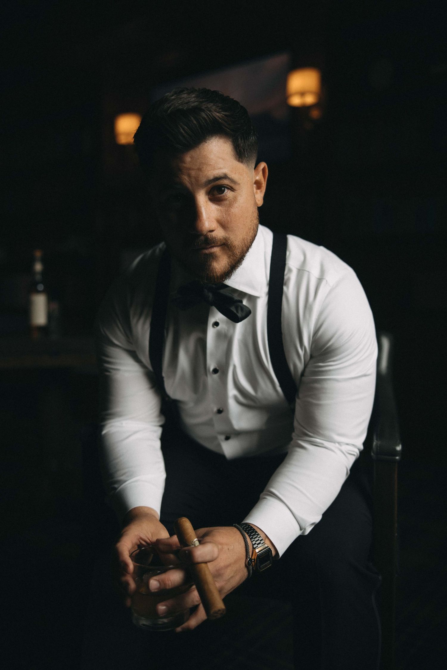 Dapper groom in white shirt, suspenders, and bow tie holds a cigar and whiskey glass in a dark setting.