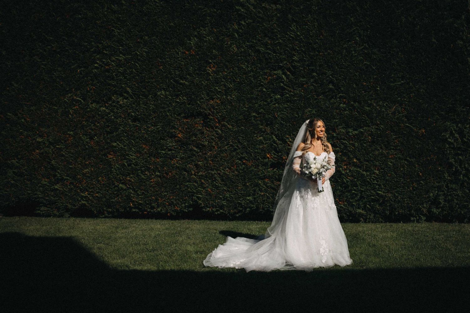 Elegant bride in a ball gown with veil holding a bouquet standing before a dark green hedge wall.