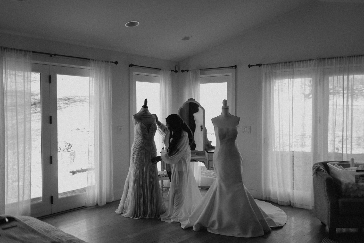 Black and white photo of bride and bridesmaids adjusting wedding gowns on mannequins by bright windows.