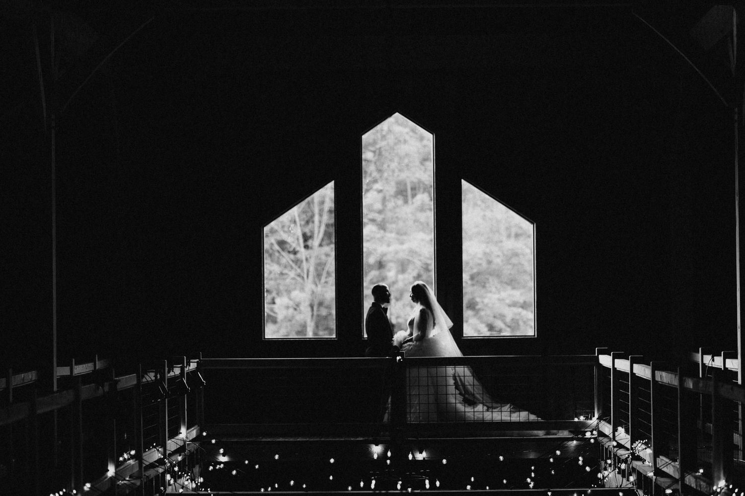 Silhouette of bride and groom holding hands before arched windows in dark romantic venue.
