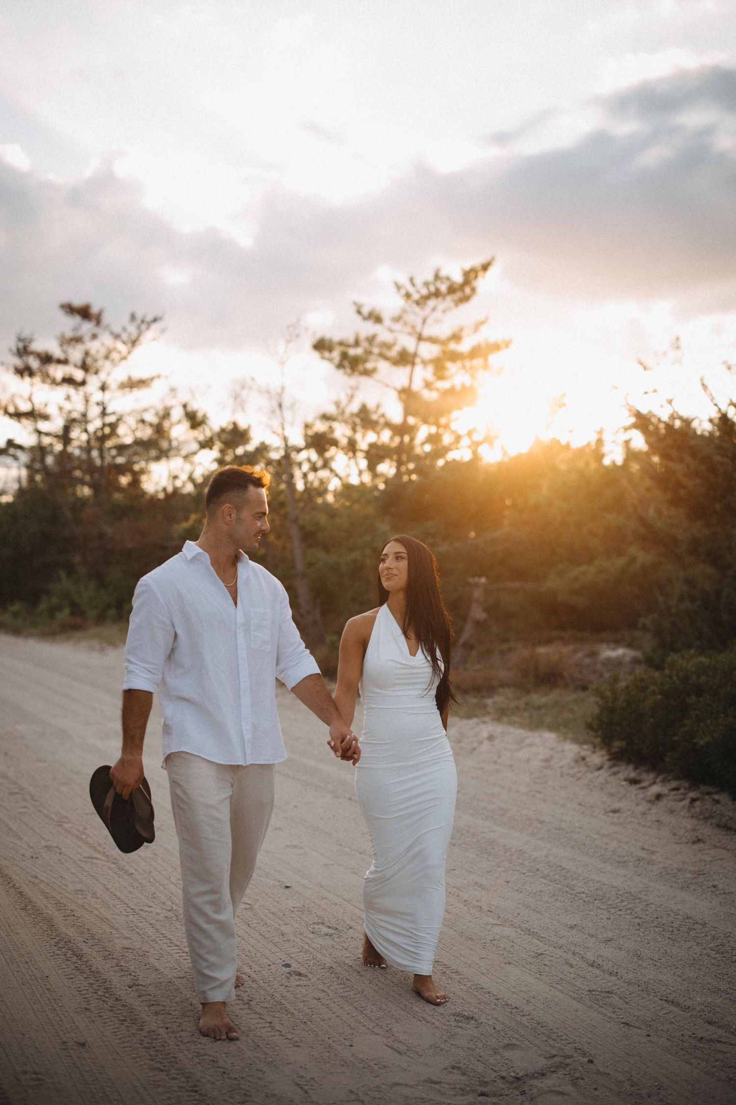 Couple in white outfits holding hands walking on sandy path at sunset.