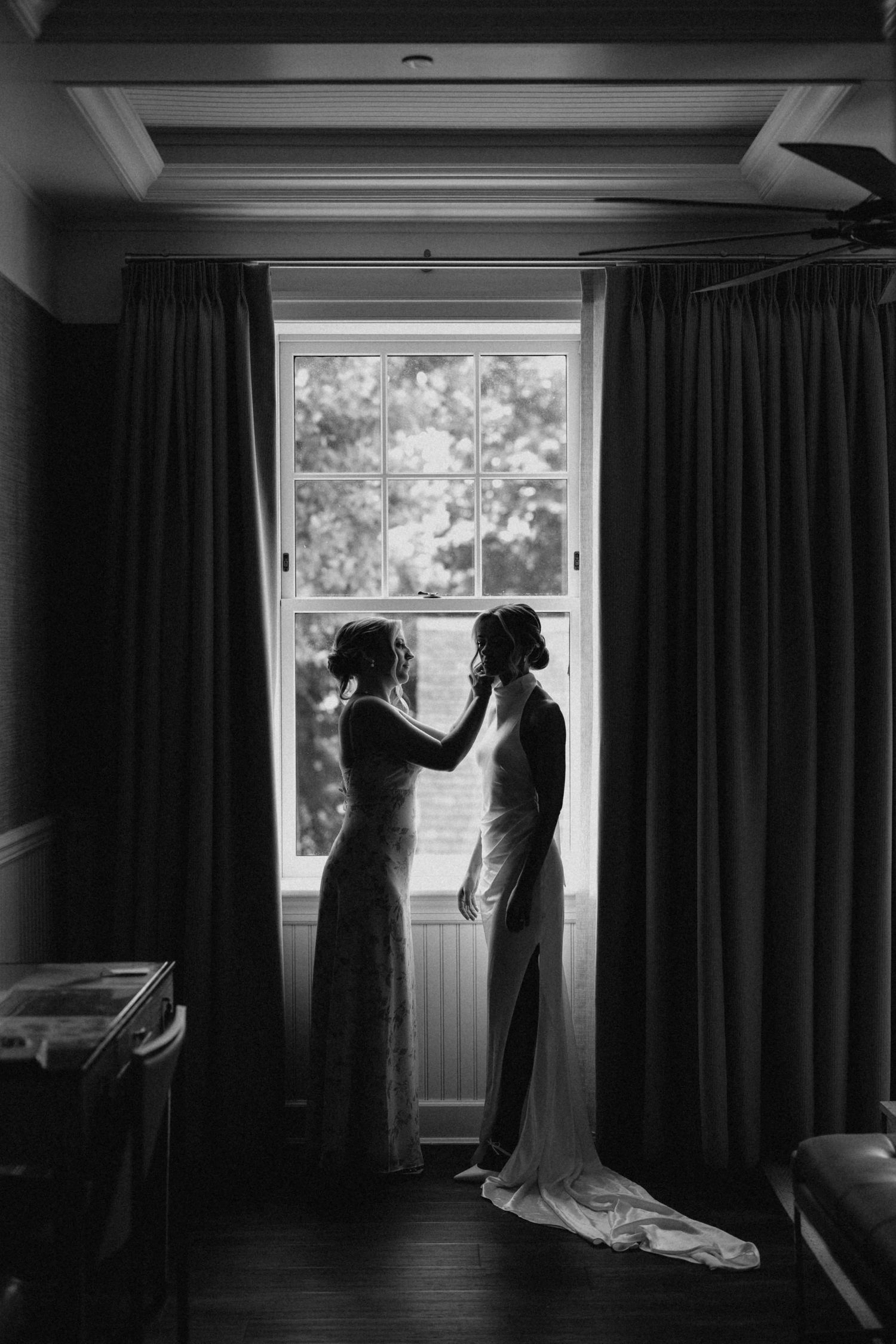 Black and white silhouette of two women in wedding attire standing by a large window.