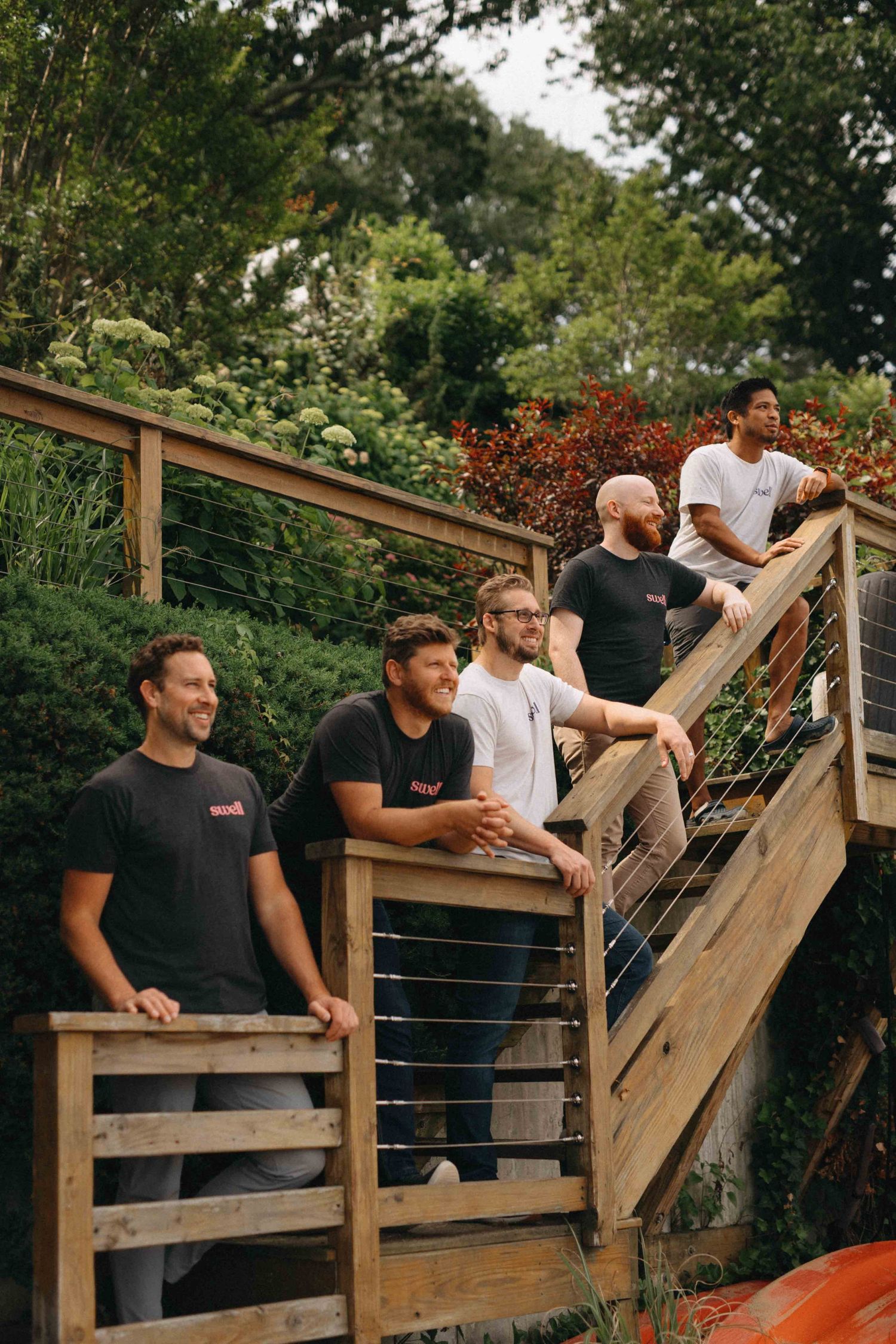 Group of five men posing on wooden outdoor staircase surrounded by lush green garden.