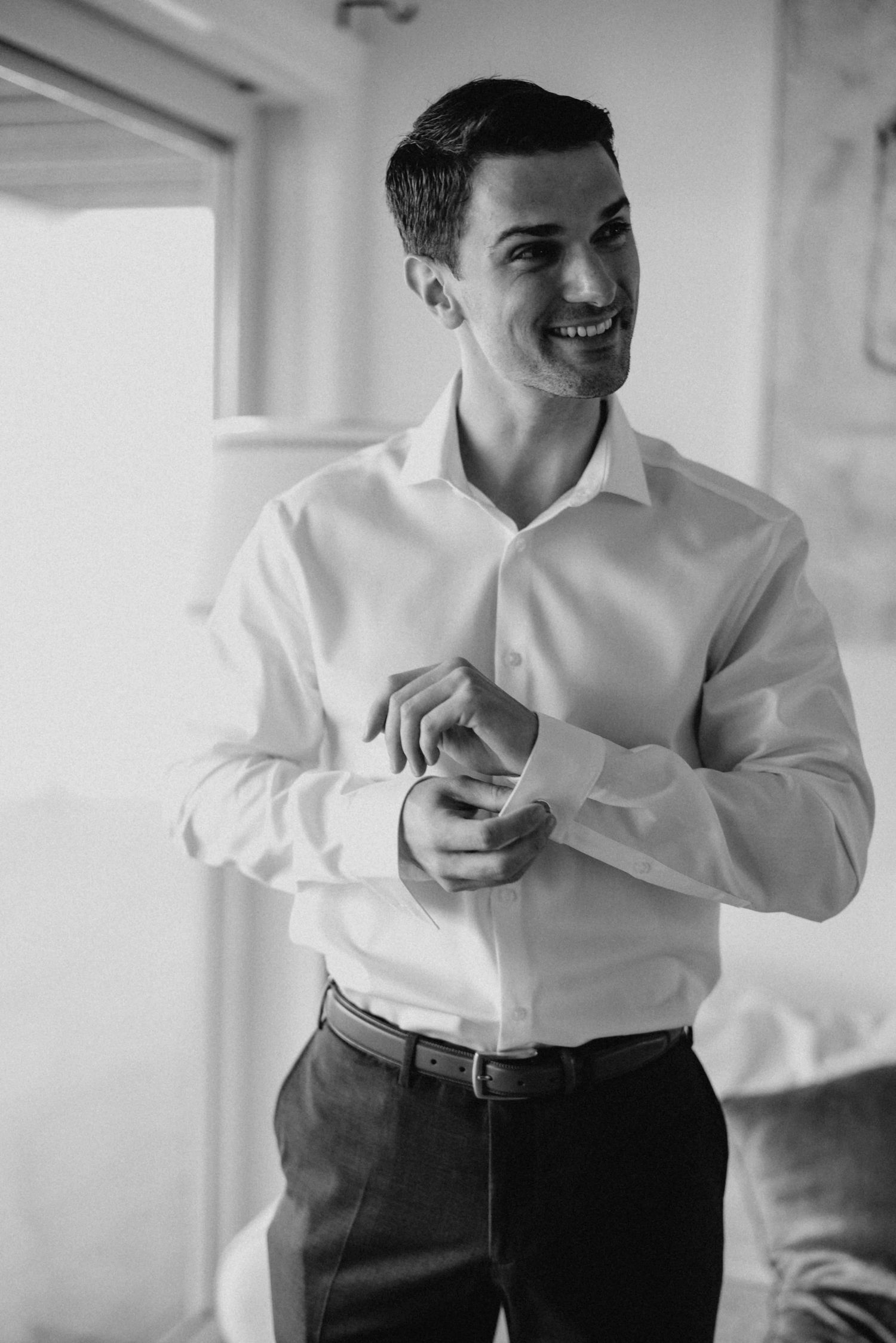Smiling groom in white dress shirt fastening cufflinks while getting ready for wedding.