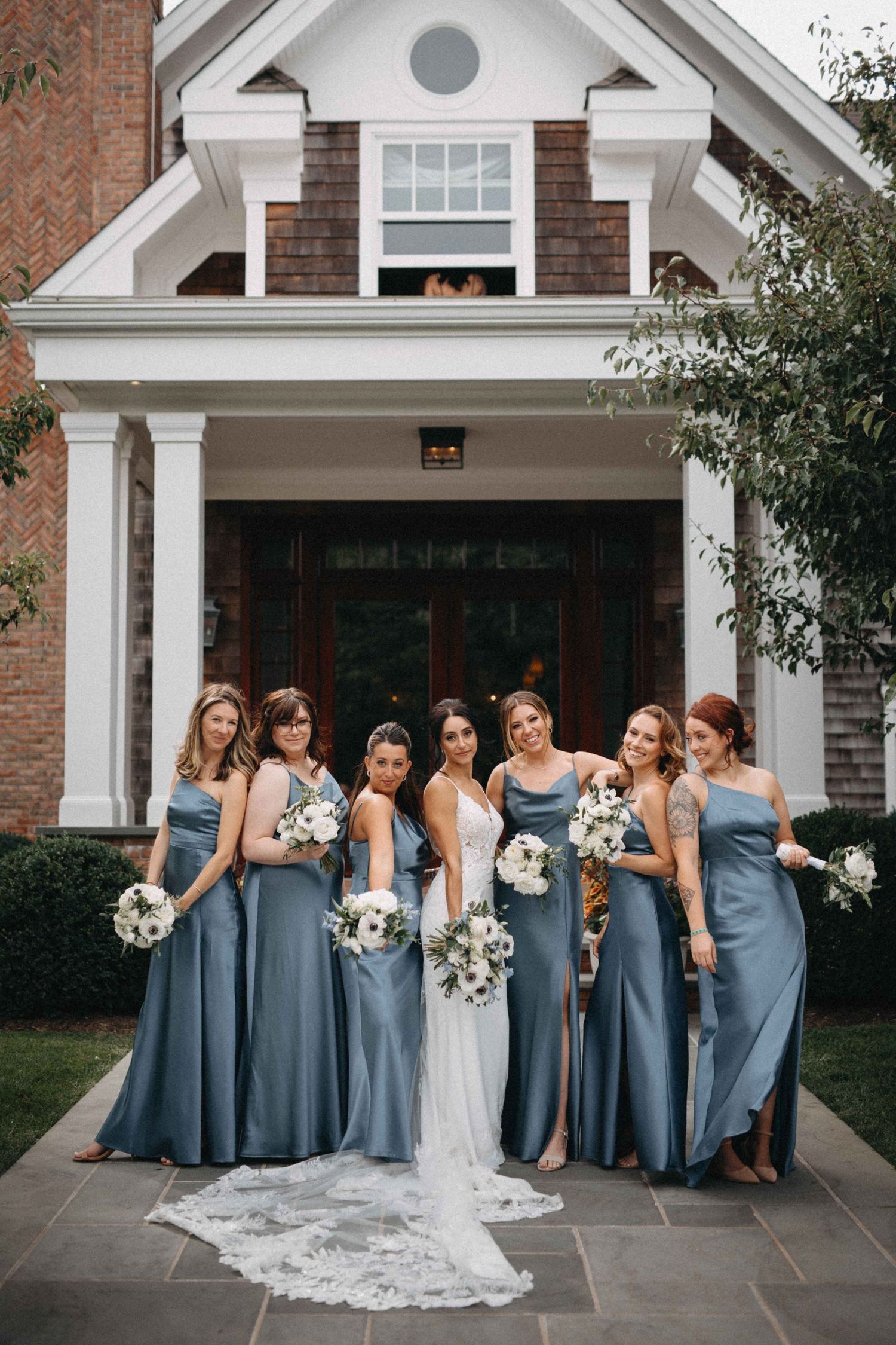 Bride in white gown with bridesmaids in dusty blue satin dresses holding white bouquets outside elegant brick home.