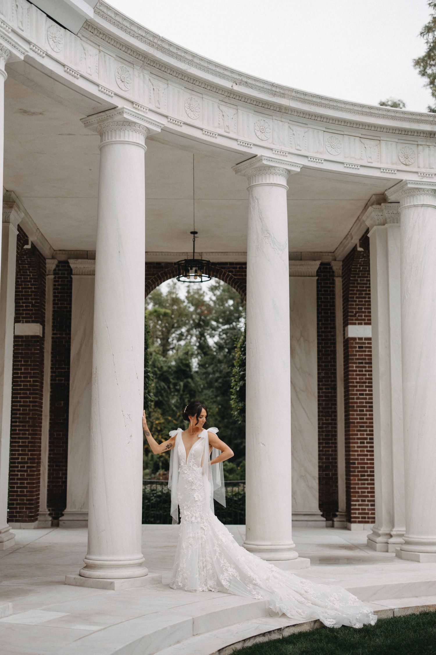 Bride in a lace mermaid gown posing gracefully beneath ornate white columns of an elegant venue.