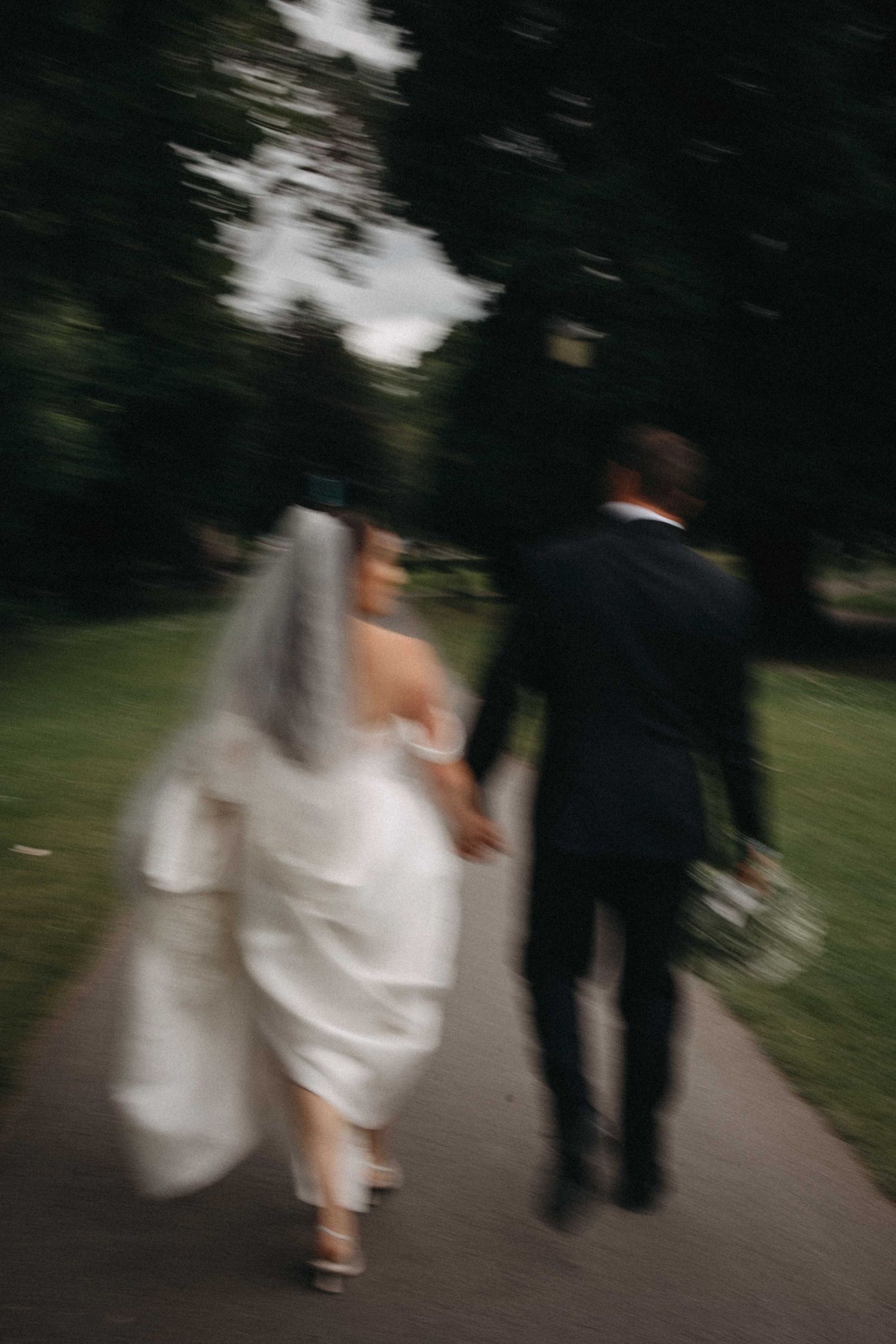 Motion-blur artistic photo of bride and groom holding hands walking along tree-lined path after wedding.