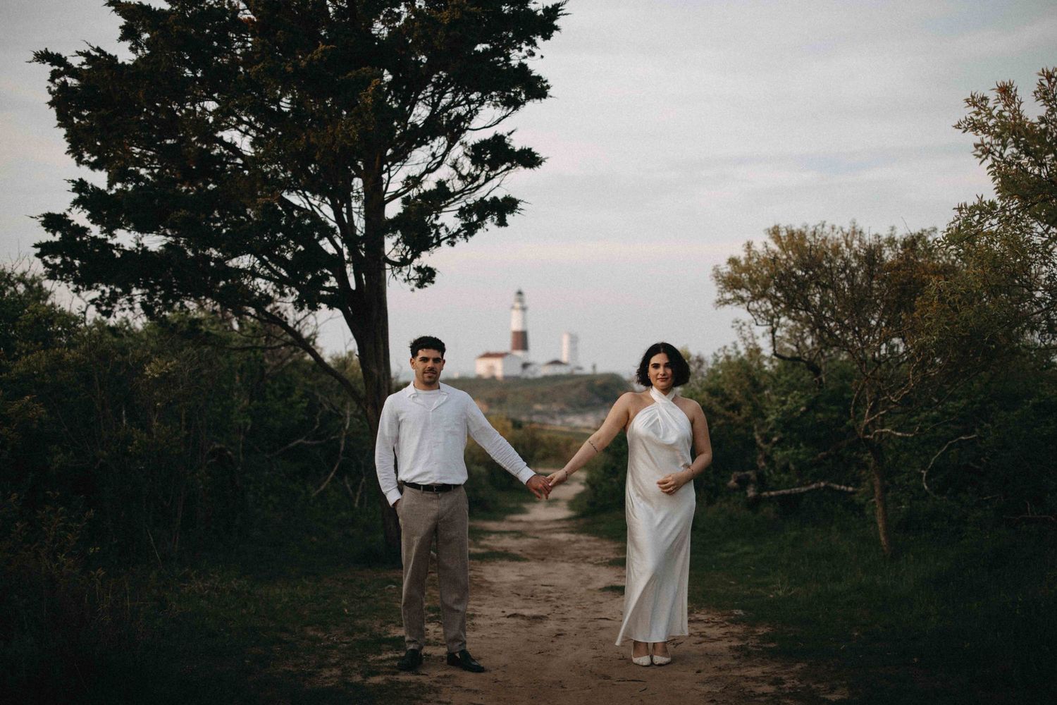 Couple holding hands on dirt path near lighthouse, woman in white halter dress, man in white shirt.