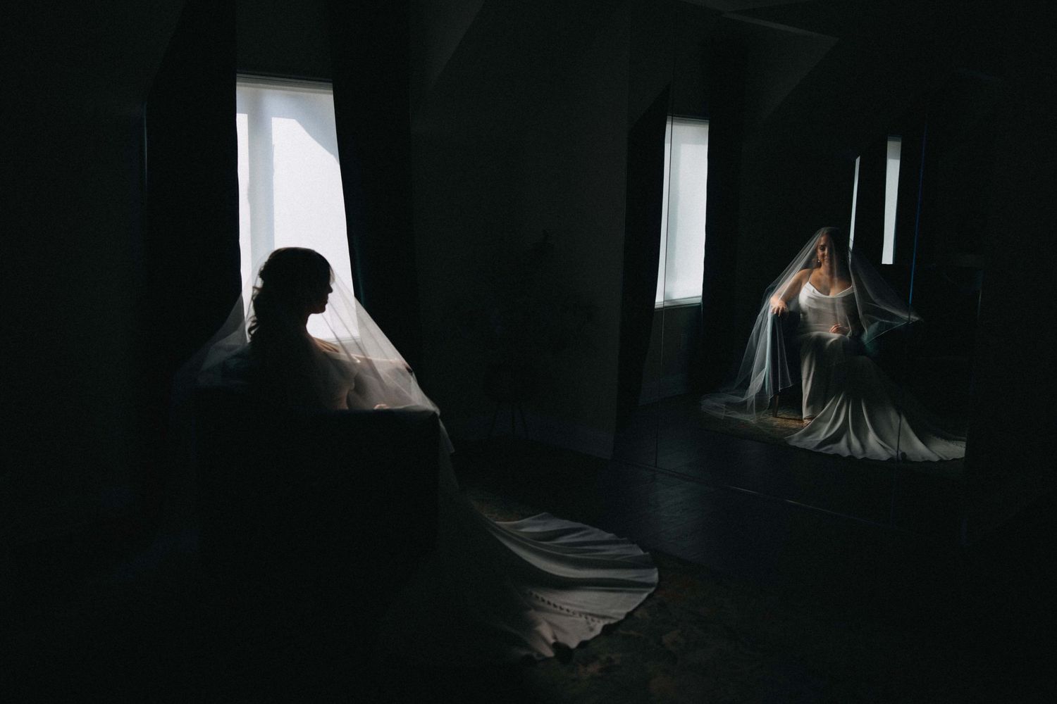 Dramatic silhouette of bride in veil seated by window in dark room.