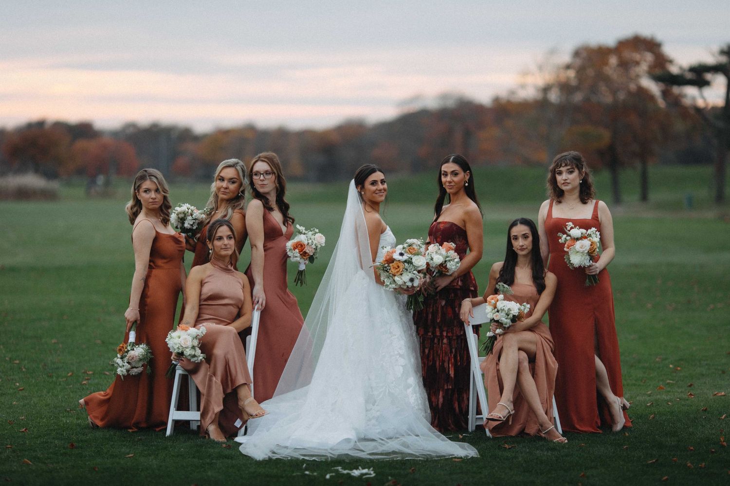 Bride with bridesmaids in rust orange gowns holding bouquets on a green lawn with fall foliage backdrop.