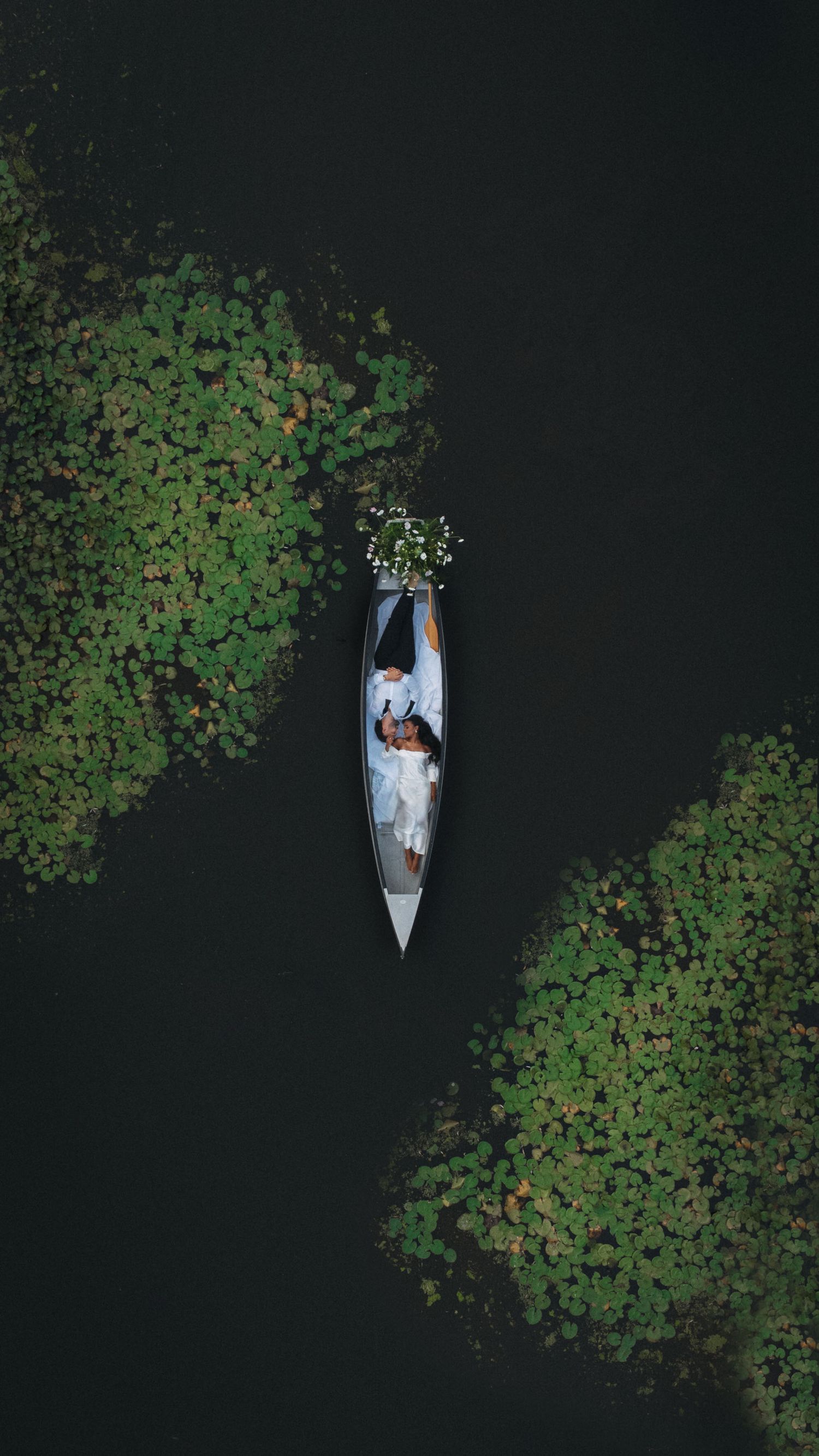 Aerial view of a couple in white lying in a narrow boat surrounded by green water lilies.