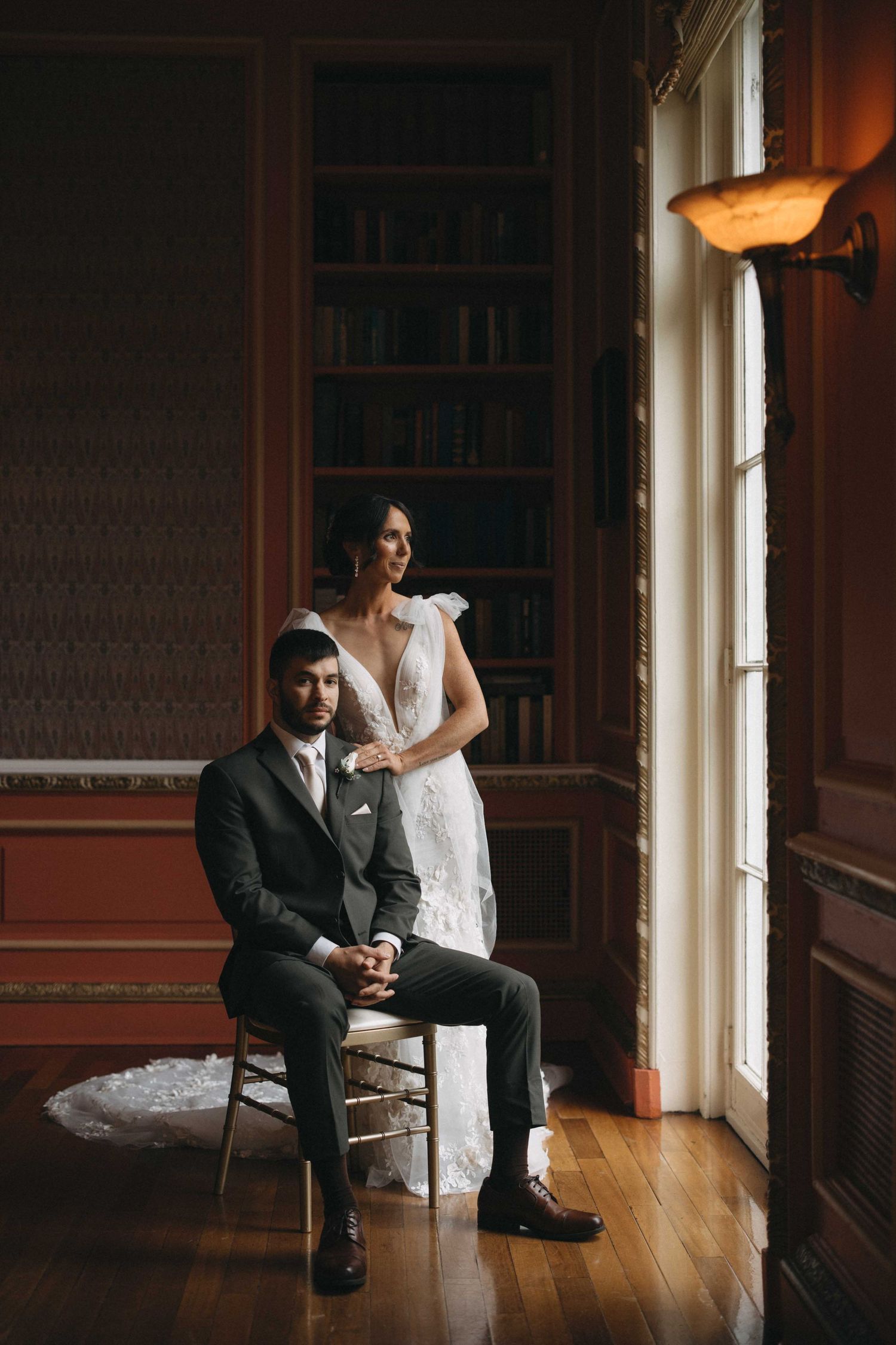 Bride and groom pose elegantly in a dark wood library room, groom seated while bride stands behind him near a tall window.