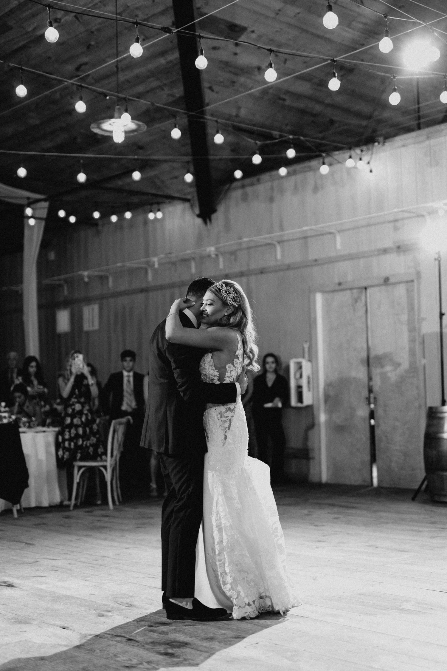 Black and white photo of bride and groom sharing first dance under string lights in barn.