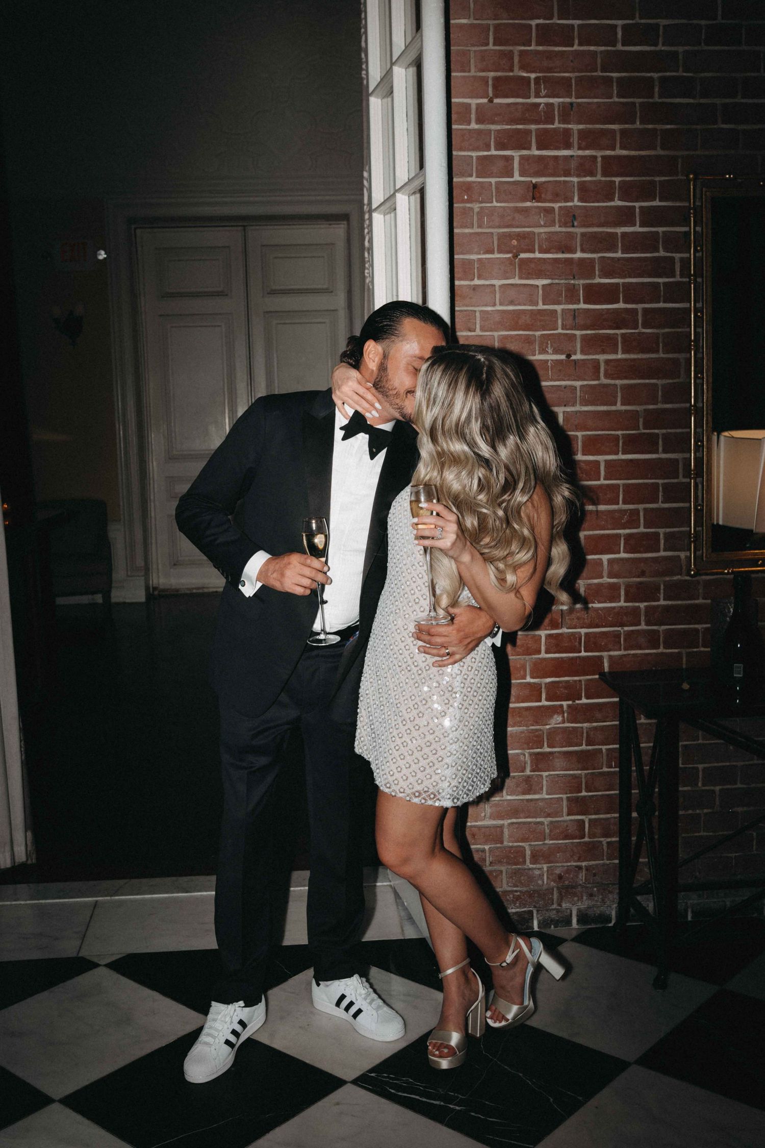 Couple sharing a kiss at wedding reception, groom in tuxedo and bride in sparkly mini dress.