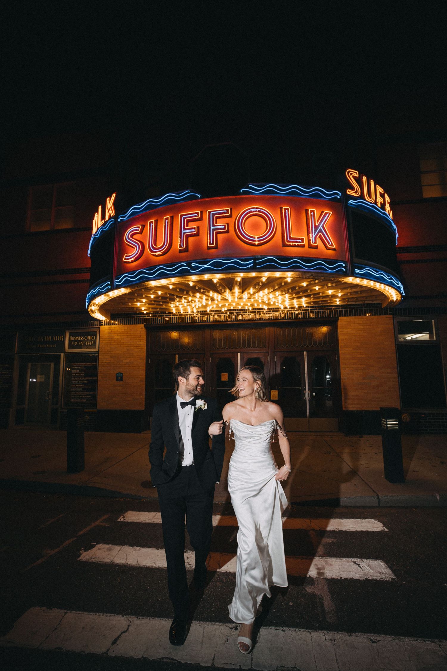Bride and groom walking under glowing Suffolk Theater marquee sign at night after wedding.