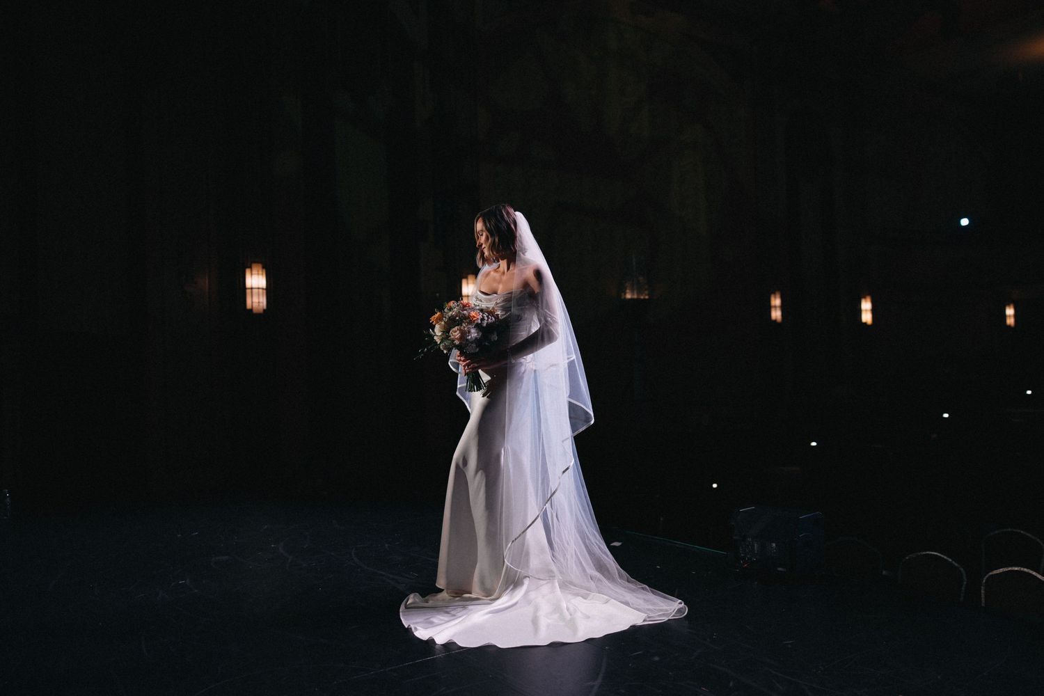 Bride in white gown and veil holds bouquet in dramatic dark outdoor wedding venue with warm ambient lighting.