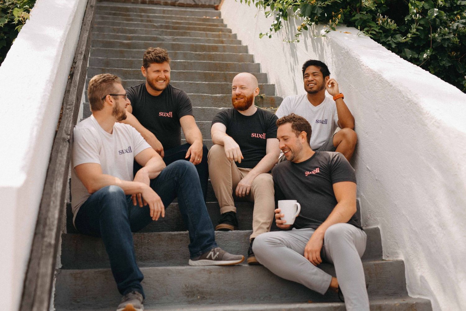 Five men sitting and smiling together on outdoor concrete steps in casual team photo.
