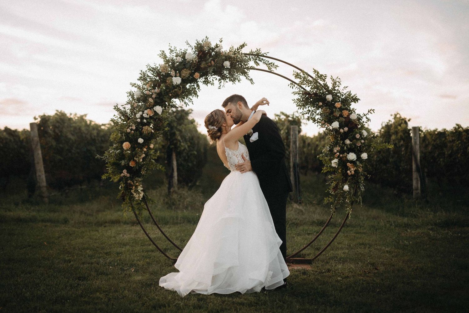 Bride and groom share a kiss under a floral circular arch in a romantic vineyard wedding.