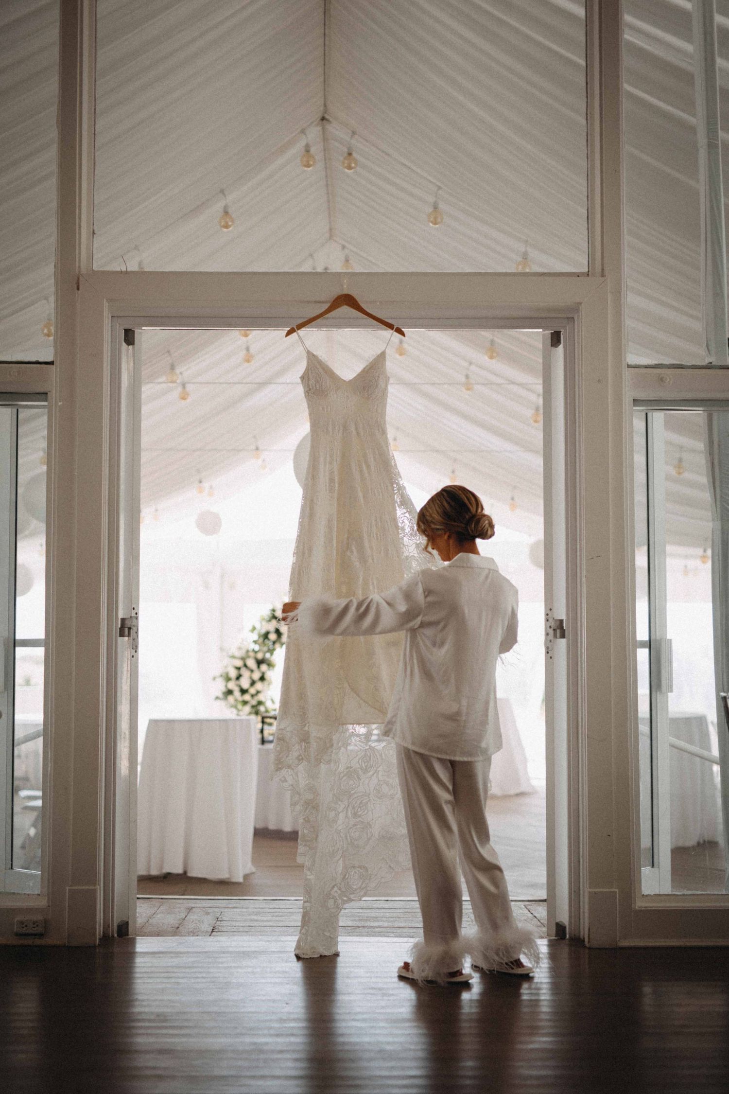 Bride in white pajamas admiring her lace wedding gown hanging in a bright venue.