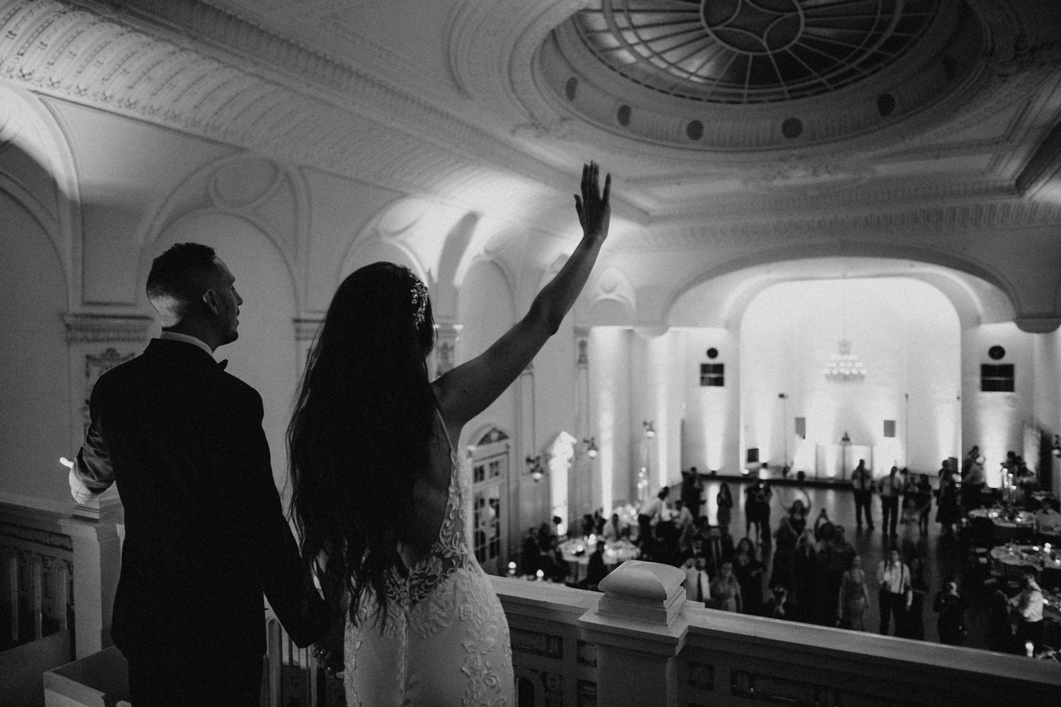 Bride and groom overlook elegant wedding reception hall from balcony in stunning black and white photo.