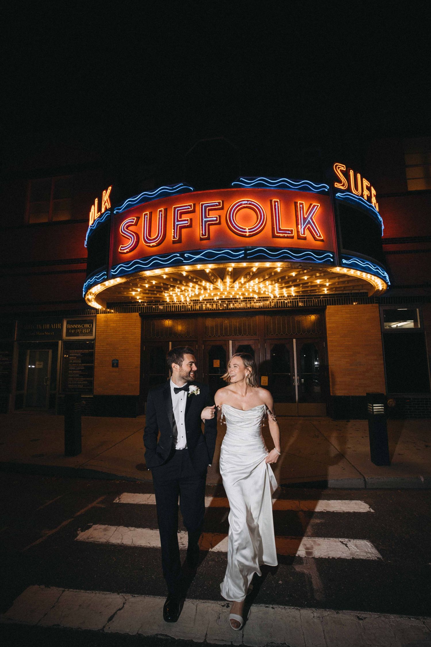 Newlyweds walk under glowing Suffolk Theater marquee sign on city street at night.