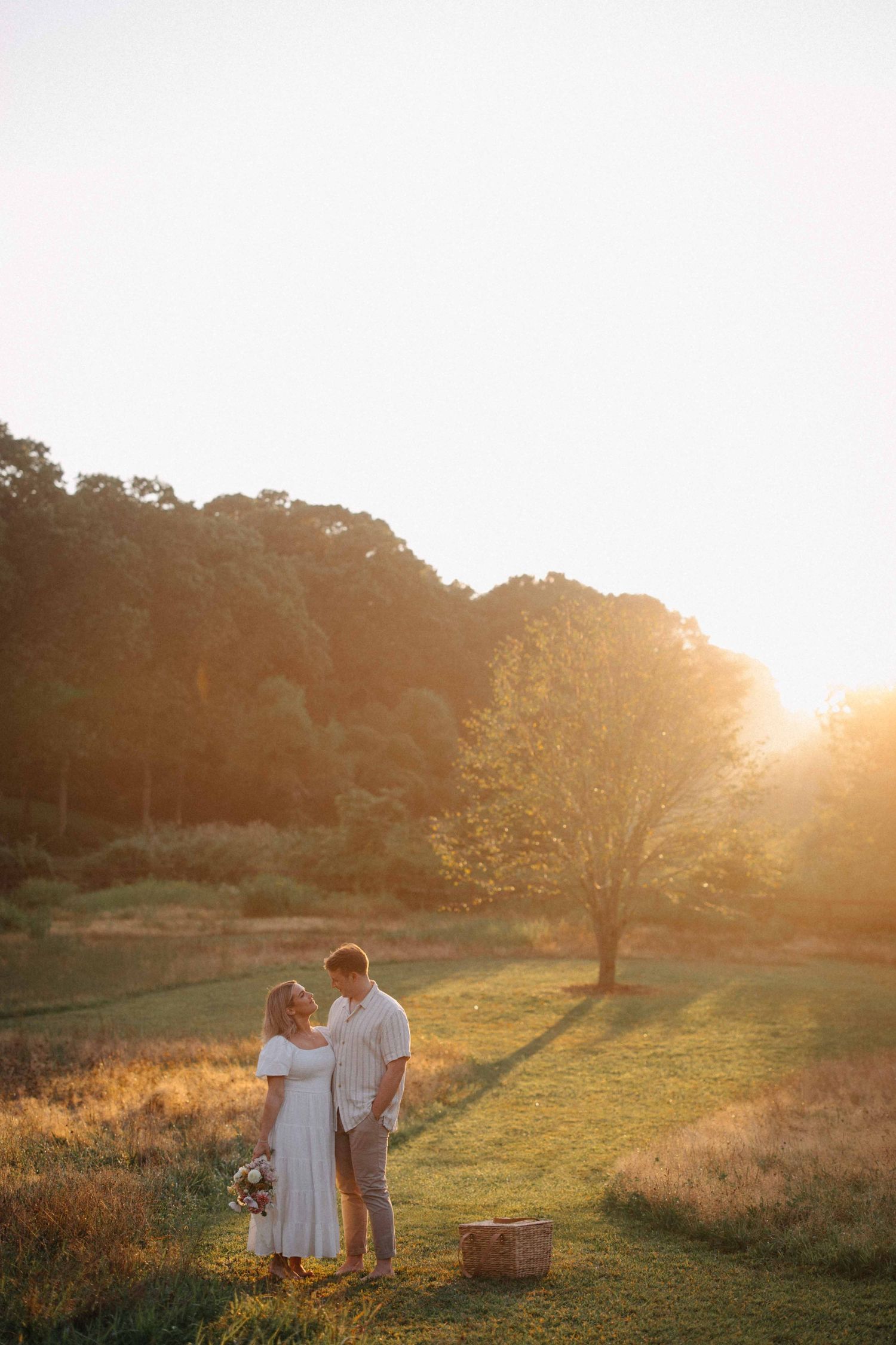Couple standing face to face in a glowing sunset meadow, woman holding a bouquet near a picnic basket.