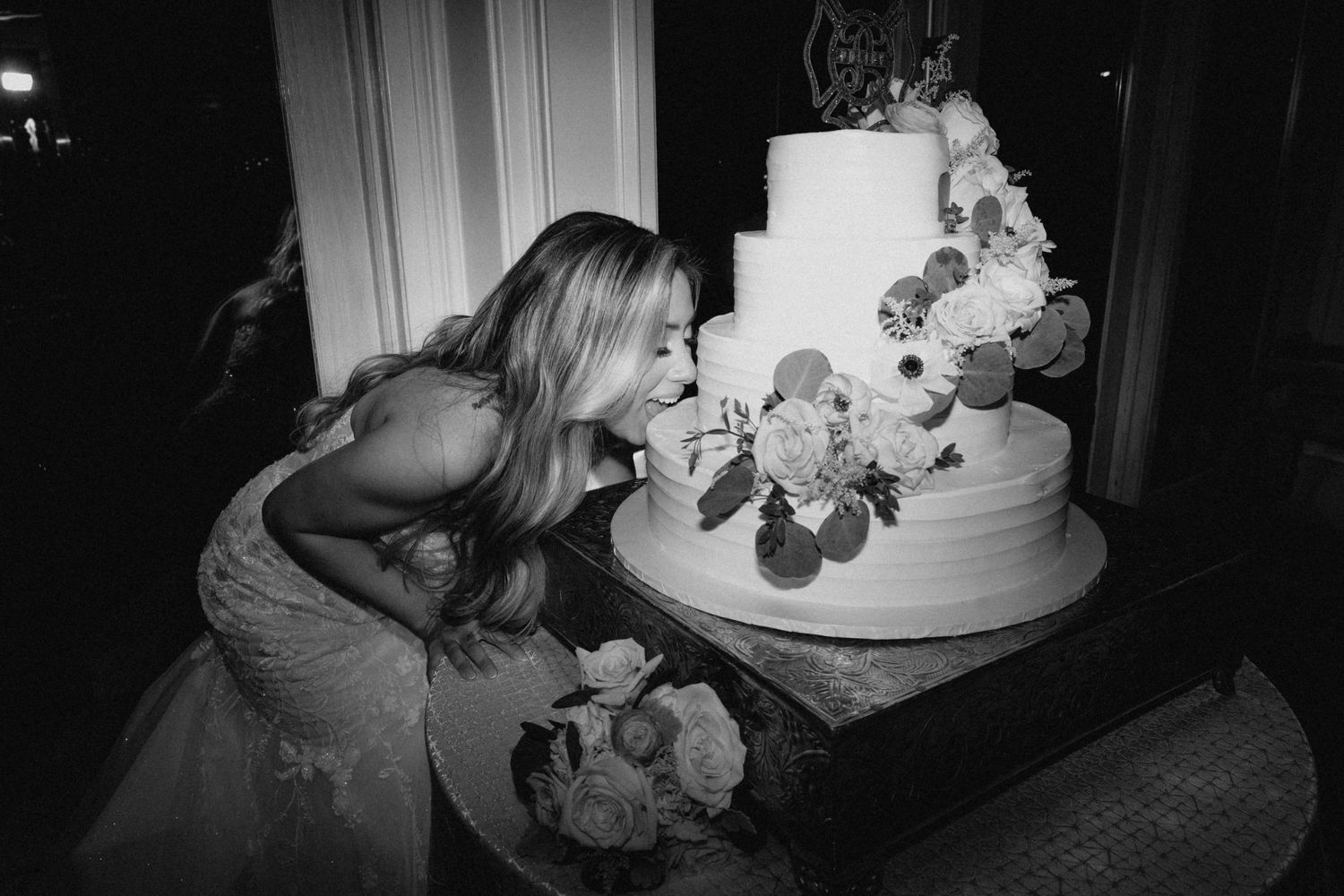 Bride in lace wedding gown leans in to taste a three-tier white floral wedding cake in black and white photo.