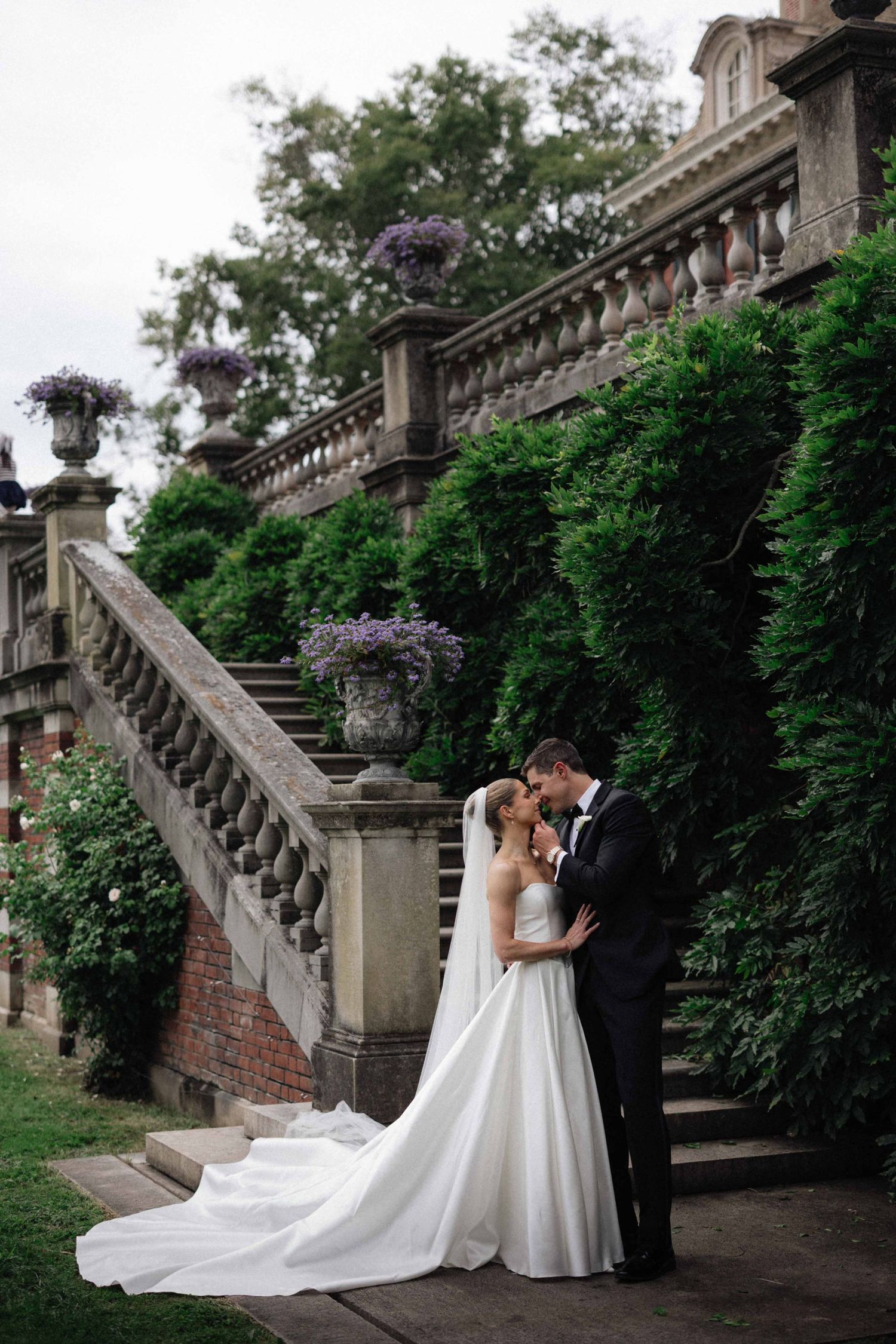 Bride and groom share romantic moment on stone staircase surrounded by lush ivy and purple flowers at elegant estate wedding.