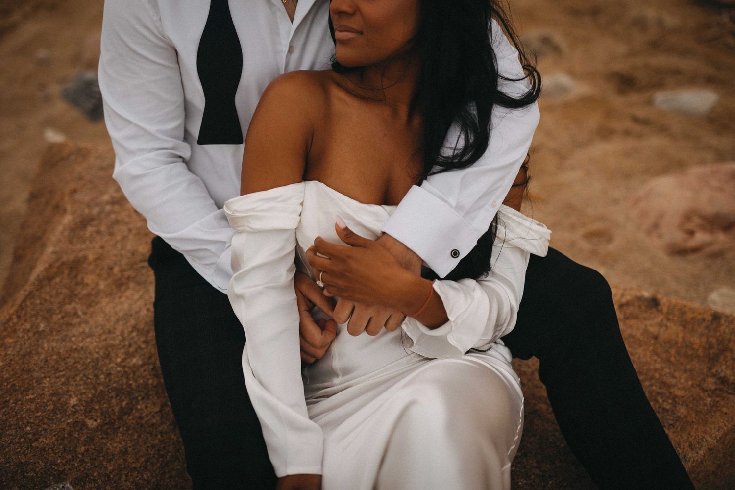 Couple in wedding attire embracing intimately on sandy beach, bride in white dress.