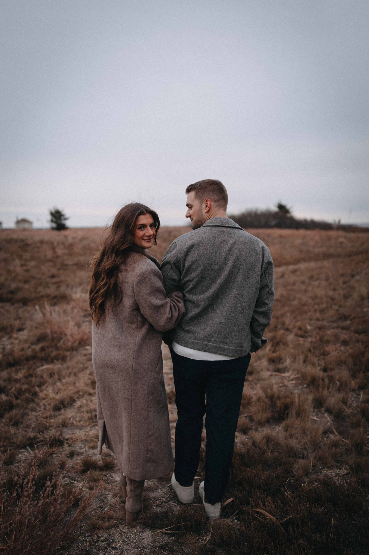 Woman smiling back at camera while walking arm-in-arm with partner through a dry autumn field.