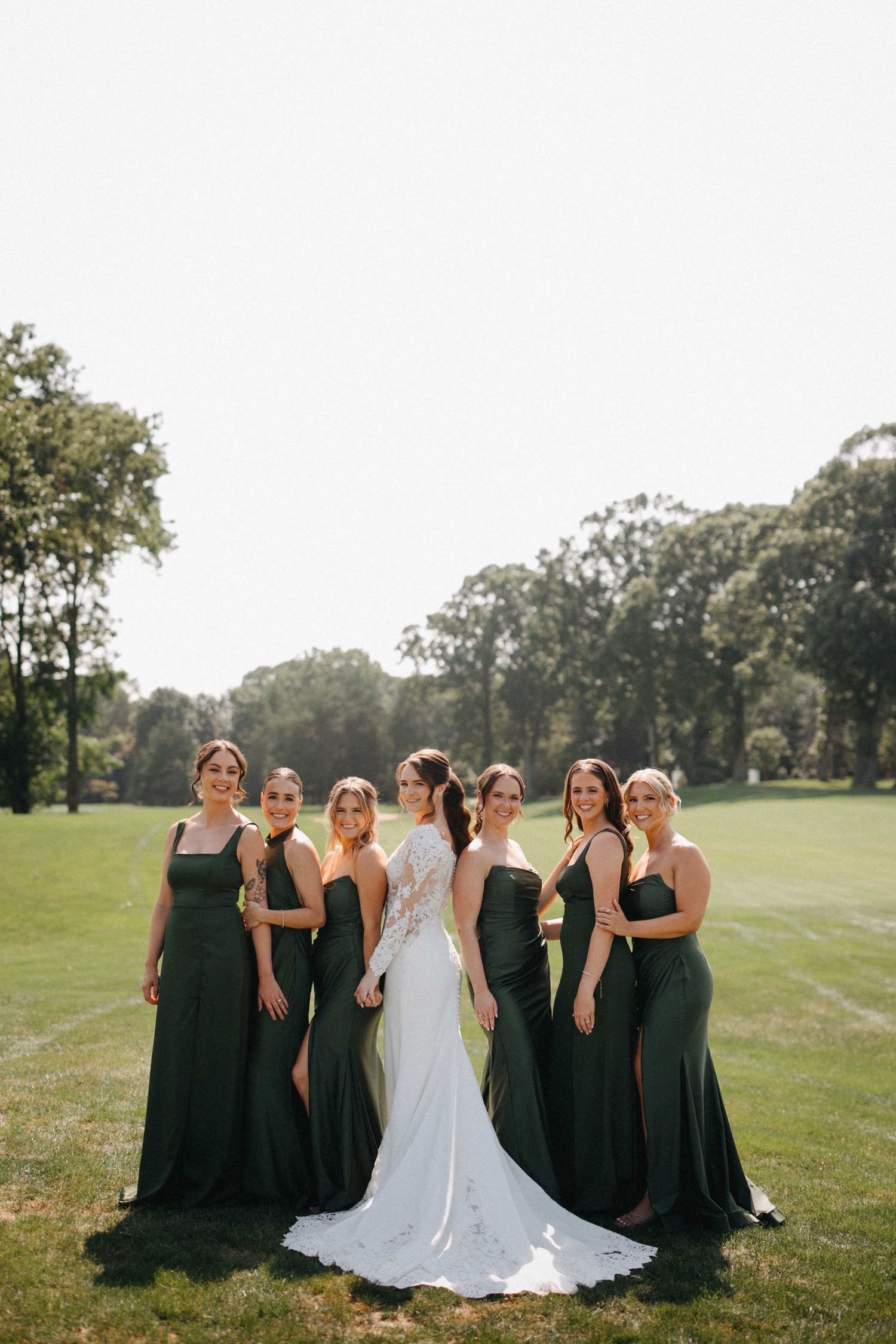 Bride in lace gown poses with six bridesmaids wearing dark green dresses on a sunny green lawn.
