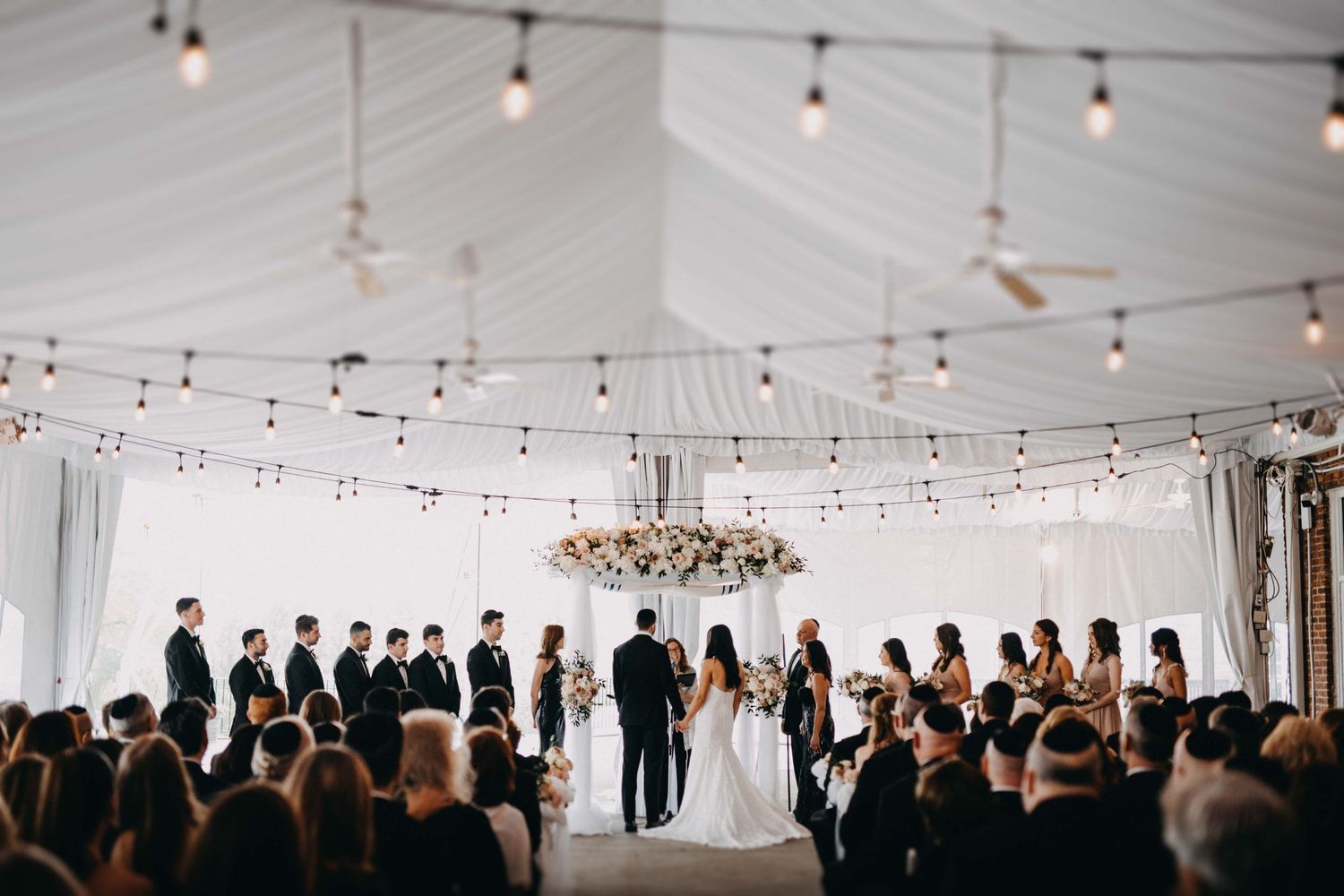 Wedding ceremony inside a white draped tent venue with string lights and a floral chandelier above the altar.