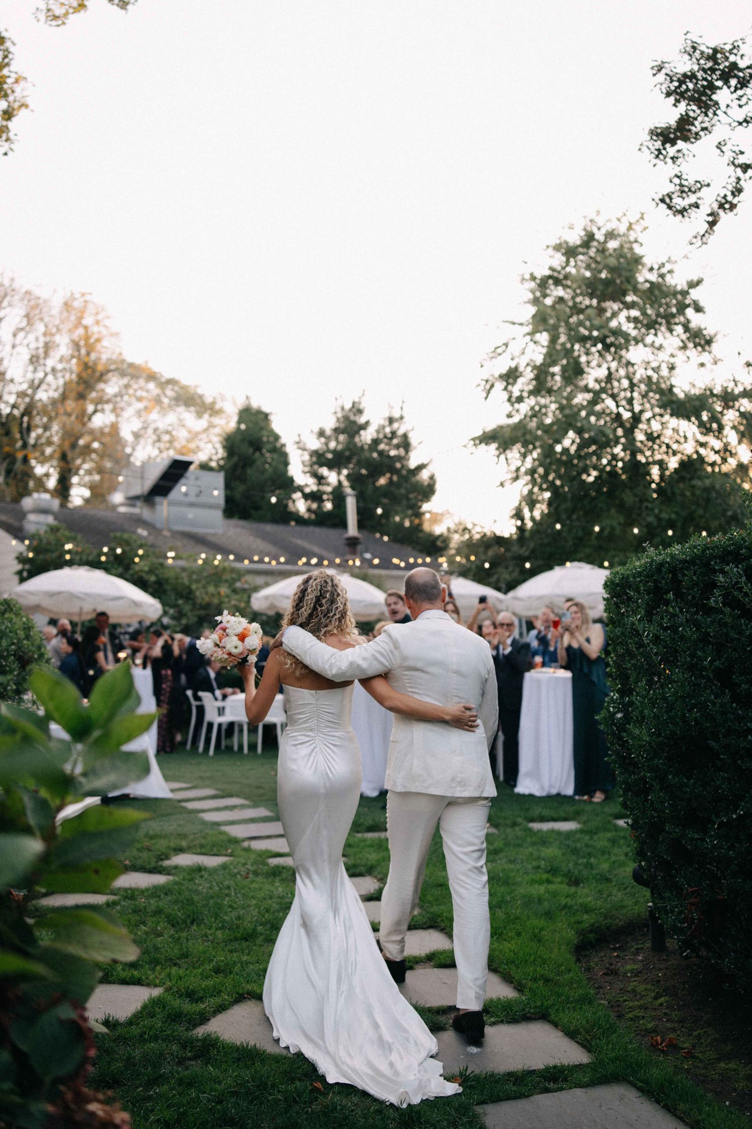 Bride in fitted white gown and groom walk arm-in-arm through an outdoor garden wedding reception at dusk.