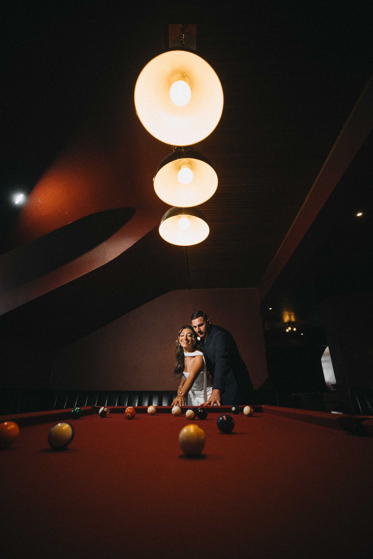 Couple poses near billiard table under warm pendant lights in dark room.
