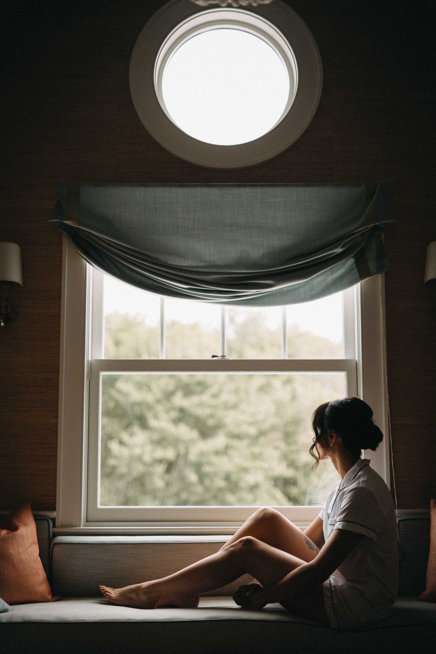 Bride sitting barefoot by a large window with round transom, gazing outside at lush greenery on her wedding day.