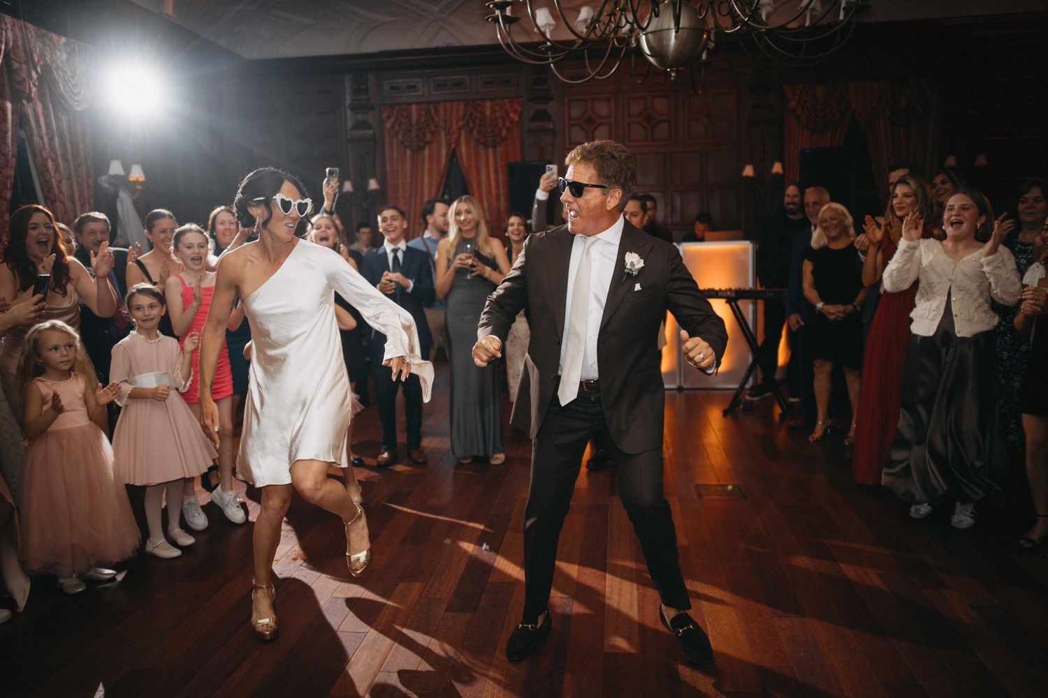 Bride and groom dance energetically in sunglasses during a fun wedding reception in a warmly lit grand ballroom.