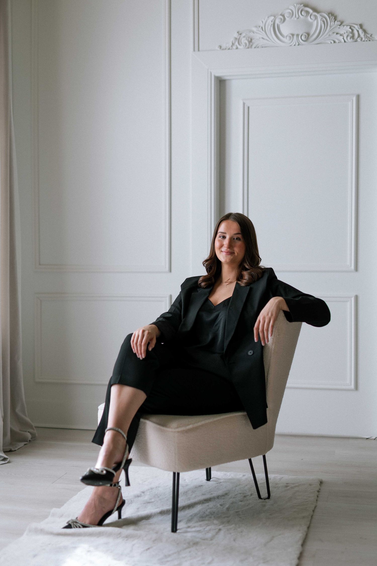 Elegant woman in black blazer and heels sitting in a beige chair against a white ornate wall interior.