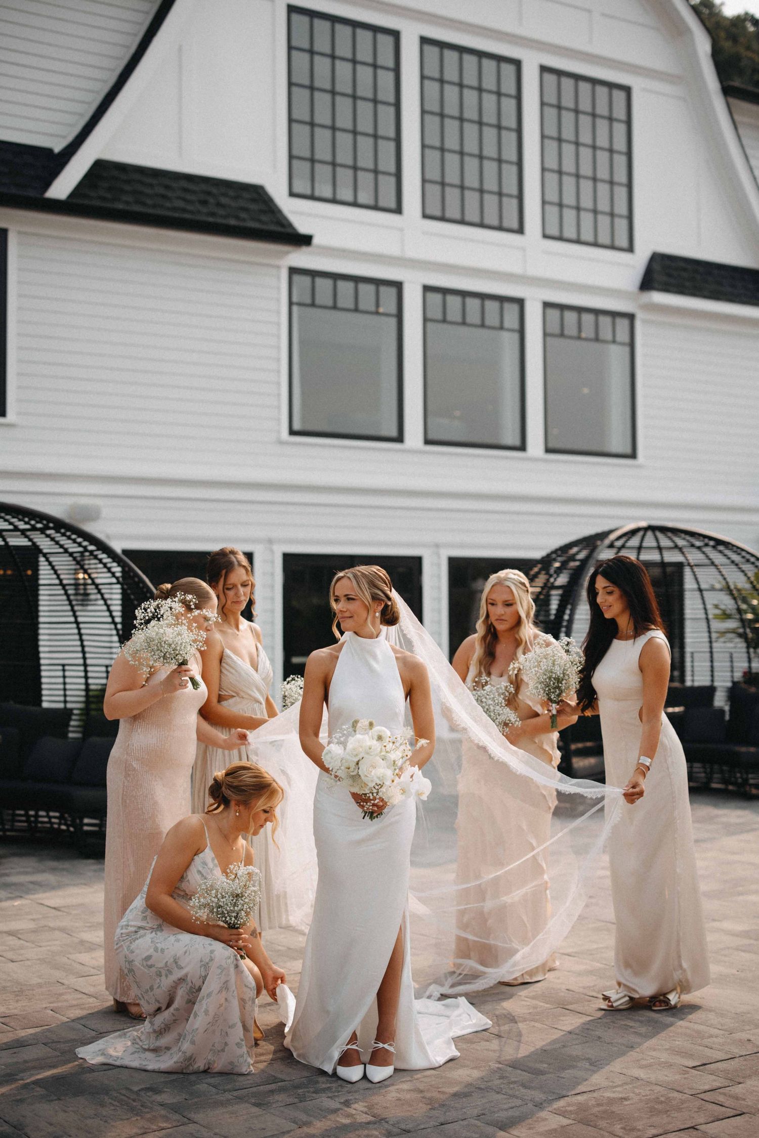 Bride in sleek white gown with veil surrounded by bridesmaids outside white barn venue.