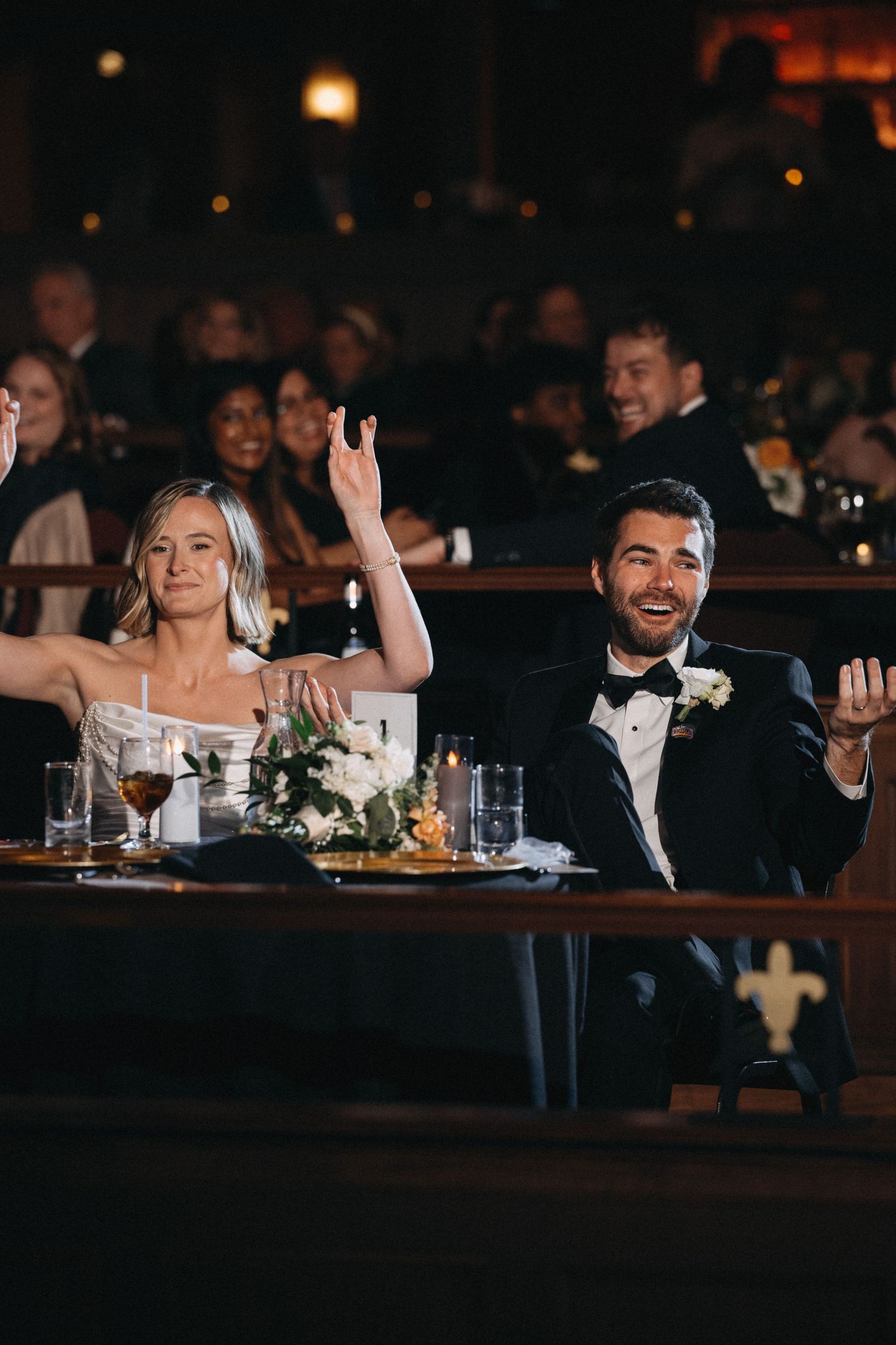 Bride and groom laugh and raise hands joyfully at their sweetheart table during reception.