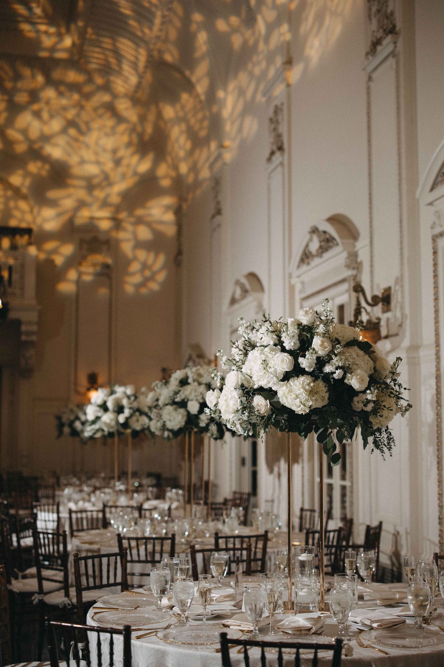 Tall white hydrangea floral centerpieces on reception tables with golden leaf light projections on ballroom walls.