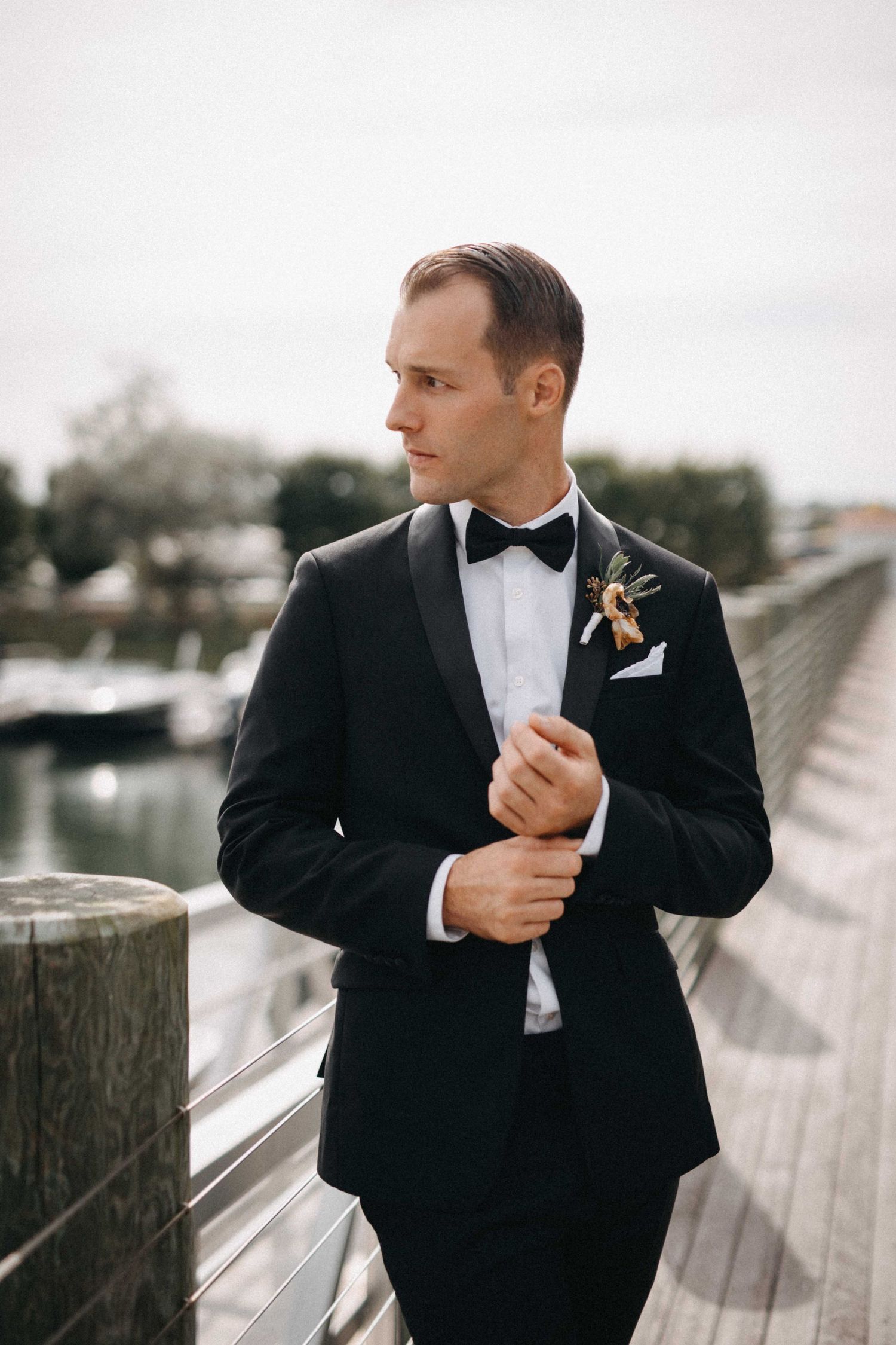 Dapper groom in black tuxedo with bow tie and boutonniere standing on wooden dock at marina.