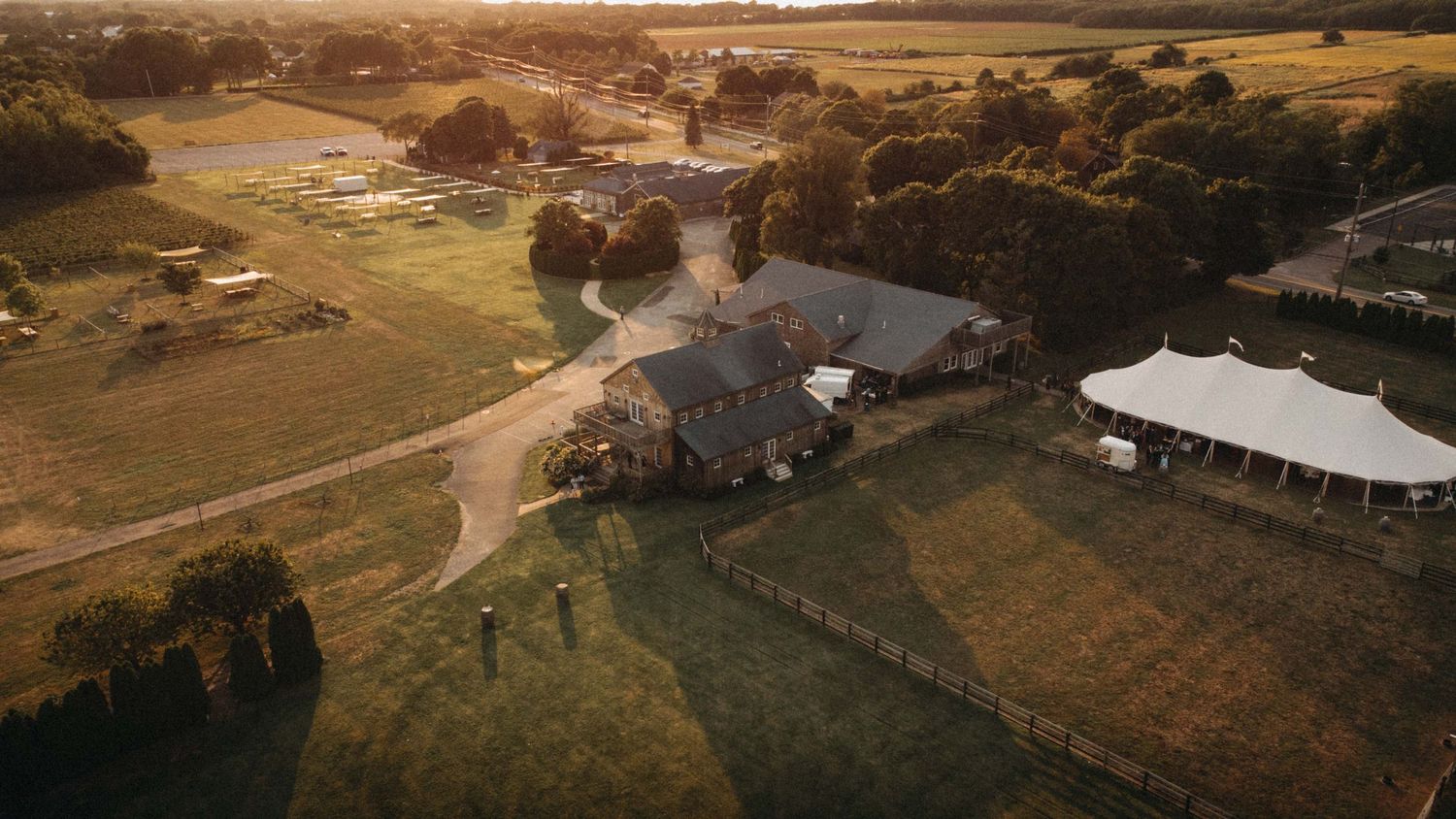 Aerial view of rustic farm wedding venue with white tent at golden hour sunset.